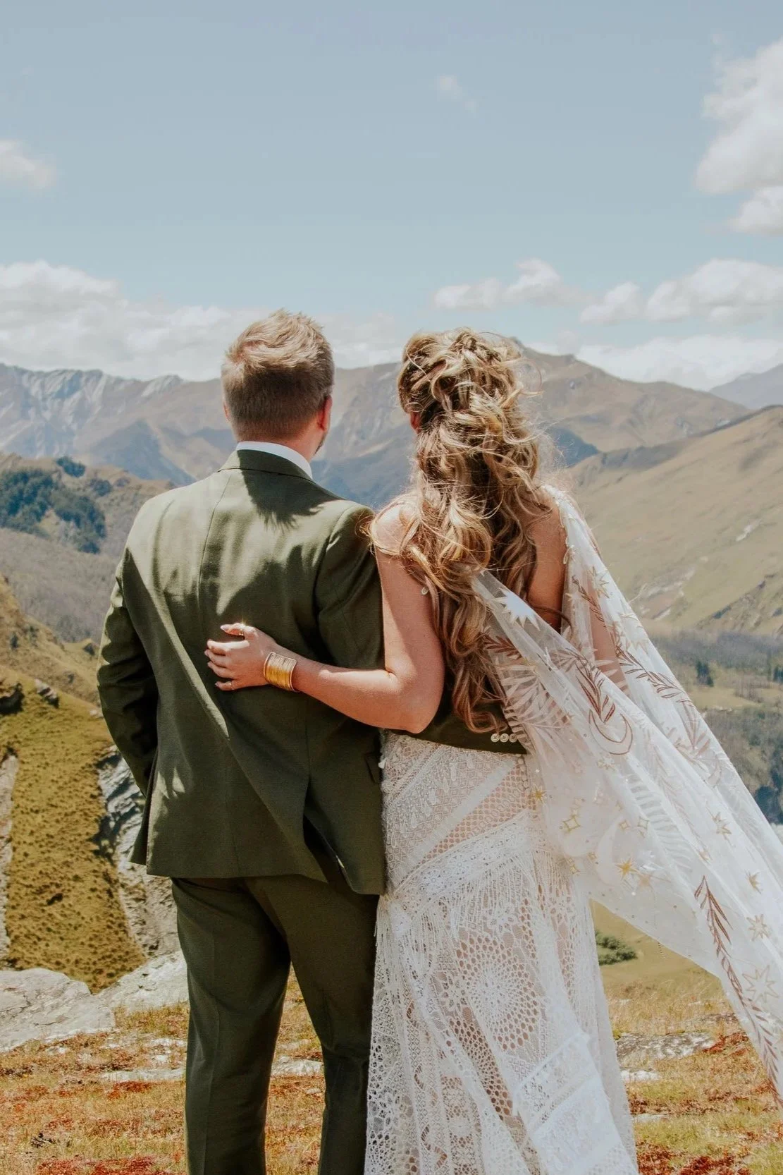 A couple dressed in wedding attire standing outdoors, with mountainous terrain in the background, facing away from the camera.