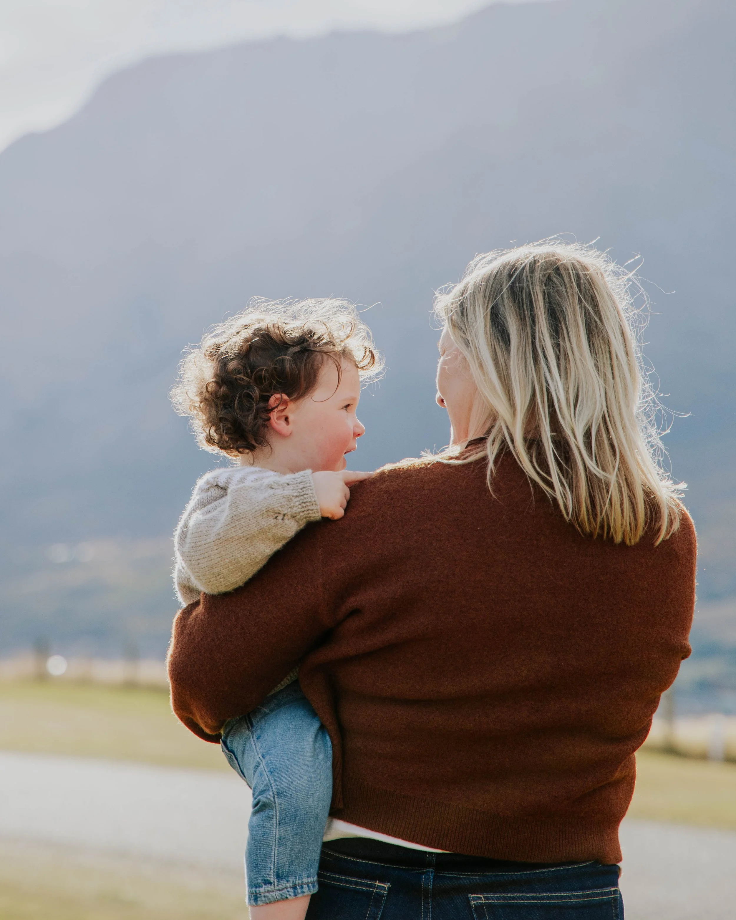 A person holding a young child with curly hair, both facing each other, outdoors in a natural setting.