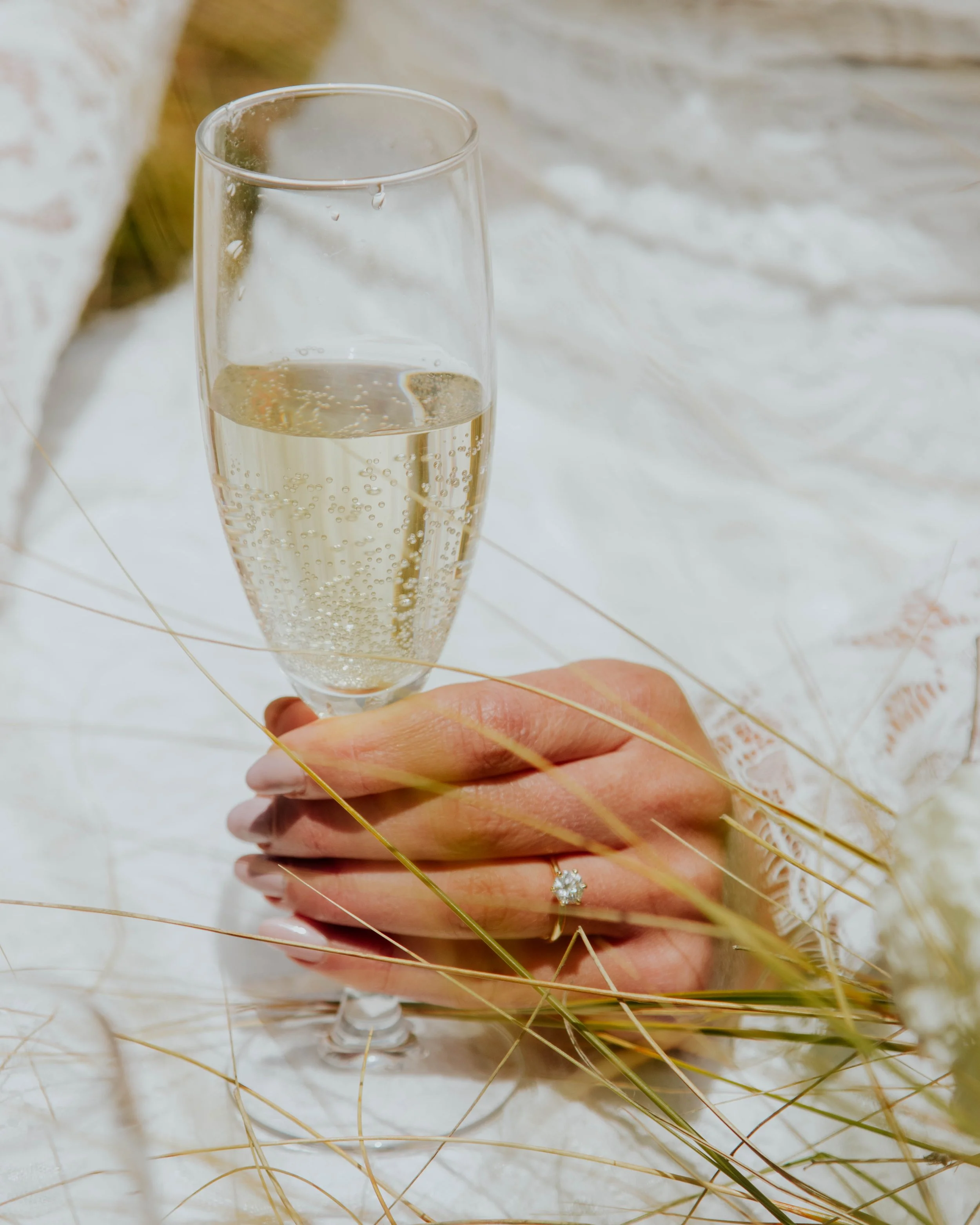 A hand holding a champagne glass filled with sparkling wine or champagne, with a diamond engagement ring on the ring finger, surrounded by dry grass and a white textured background.
