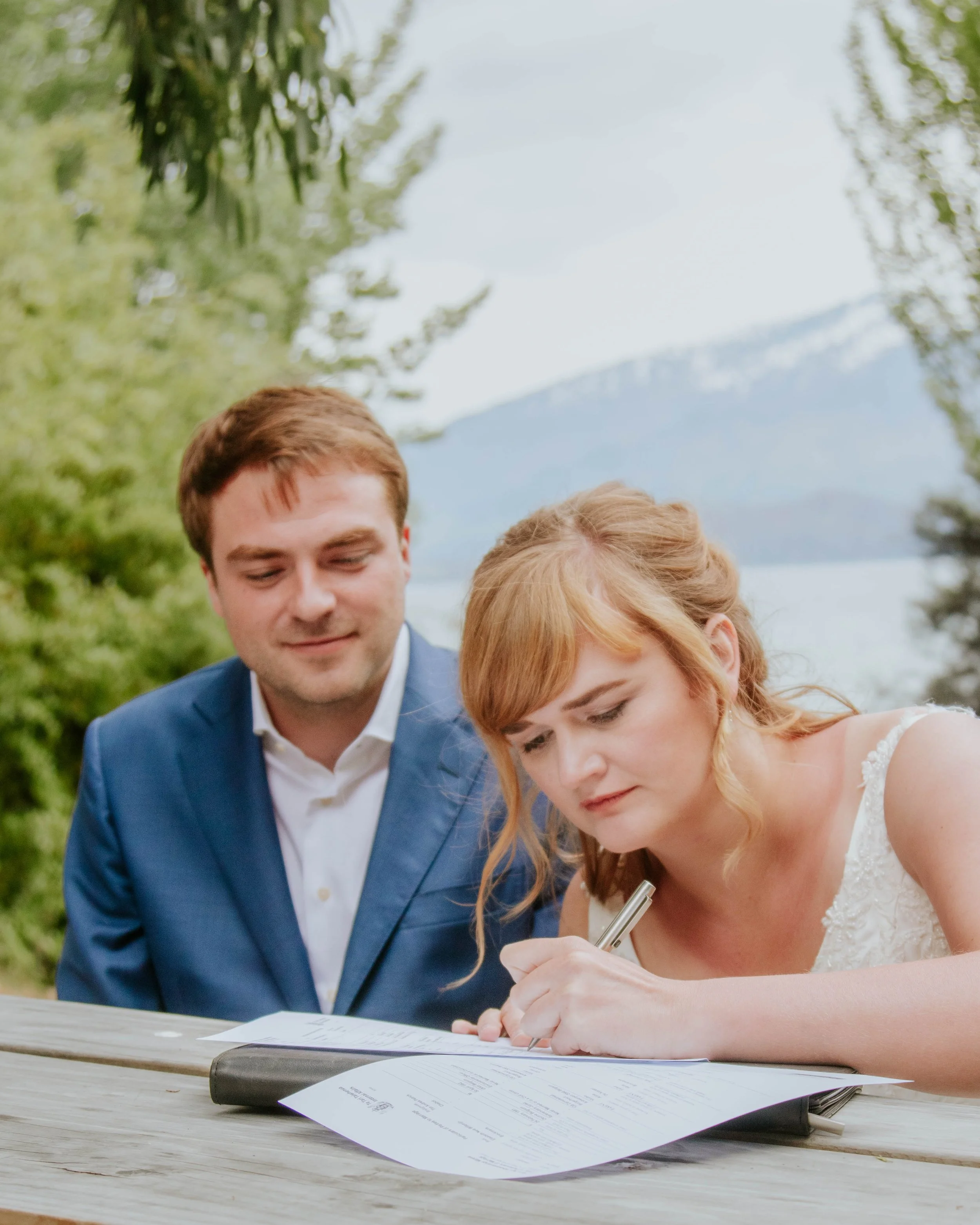 A woman in a white wedding dress signs a document on a table while a man in a blue suit looks on, outdoors near a lake with mountains in the background.