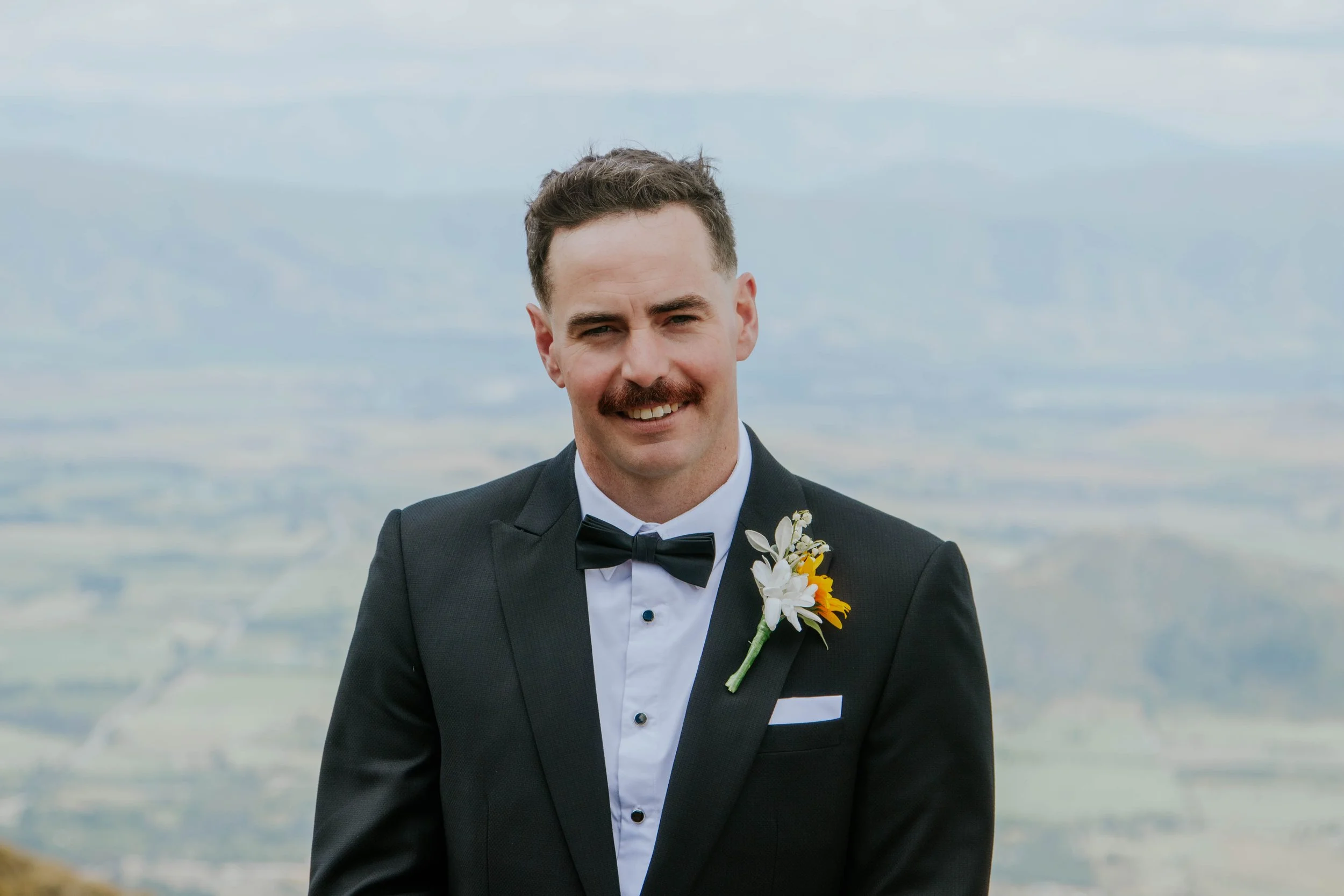 A man in a tuxedo with a boutonniere on his lapel, standing outdoors with a scenic landscape in the background, smiling.