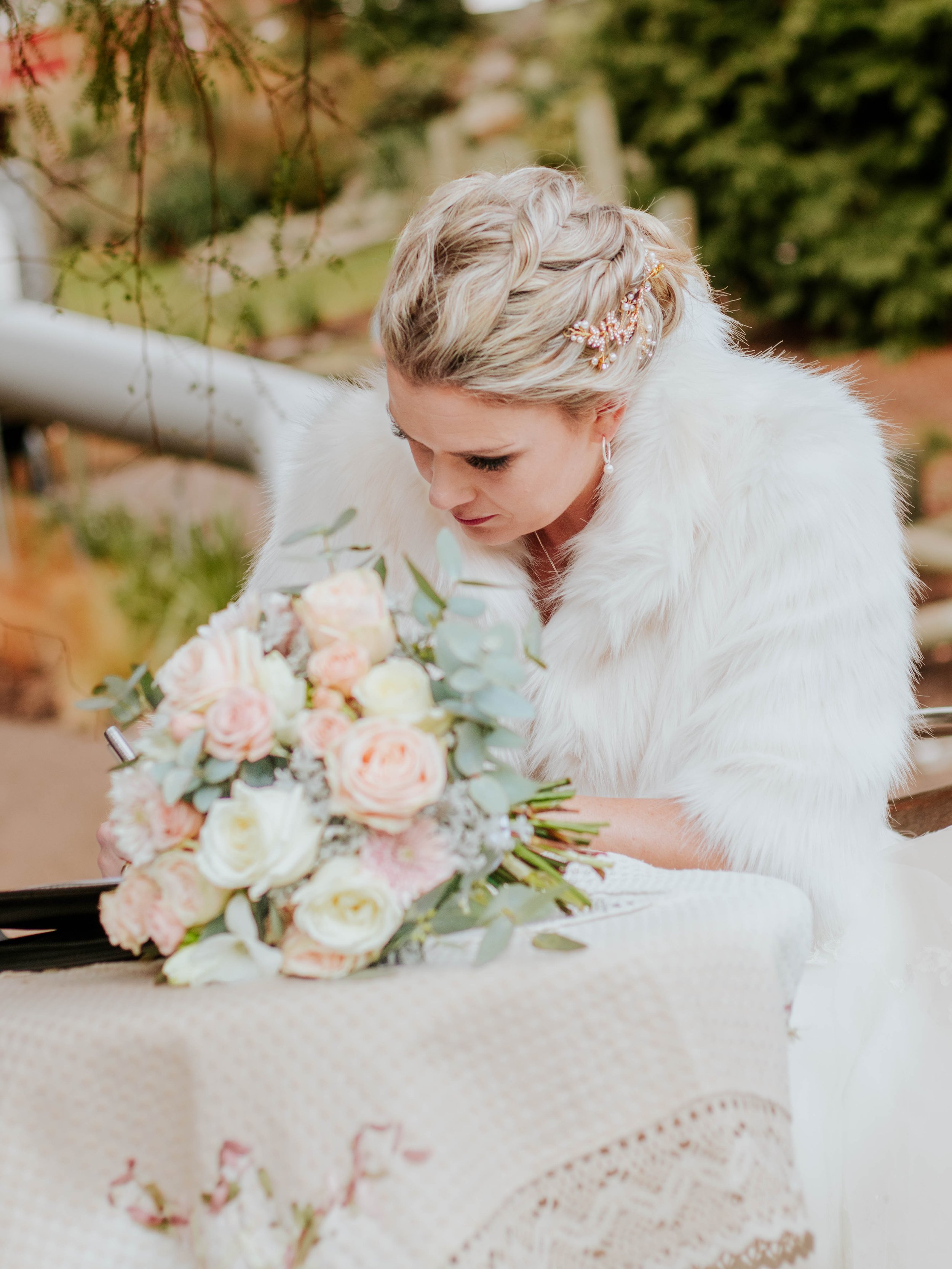 A woman with blonde hair styled with a braid and floral hair accessory, wearing a white faux fur shawl, sitting at a table with a bouquet of pink and white roses and greenery, outdoors.