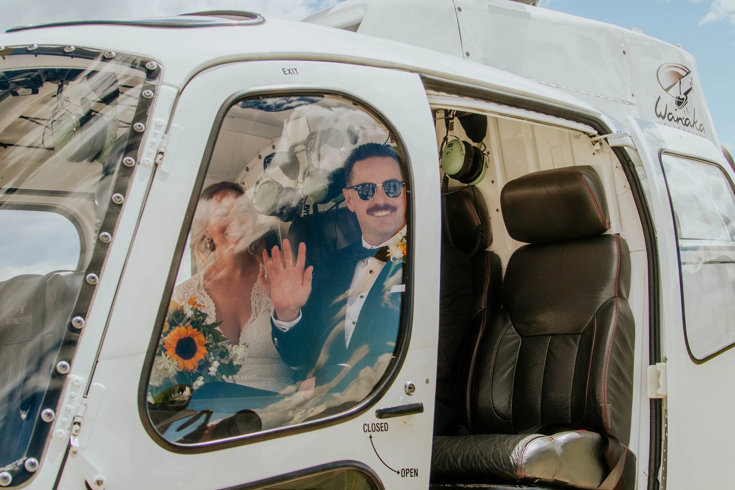 A couple dressed in wedding attire inside a helicopter, smiling and waving through the window. The man is wearing sunglasses and a tuxedo, holding a bouquet of sunflowers, while the woman has a lace dress and a floral bouquet.