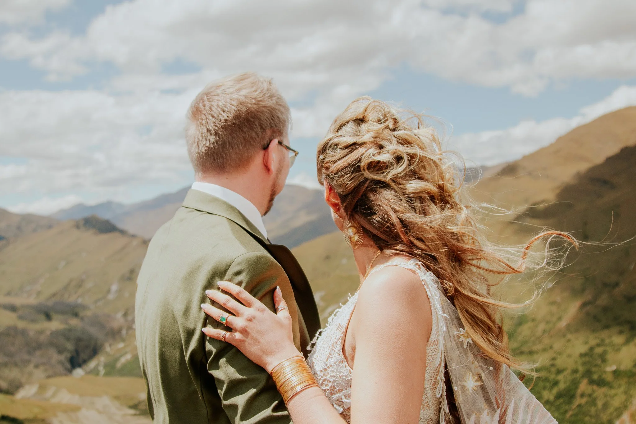 A couple stands close together outdoors with mountains in the background, dressed in wedding attire.