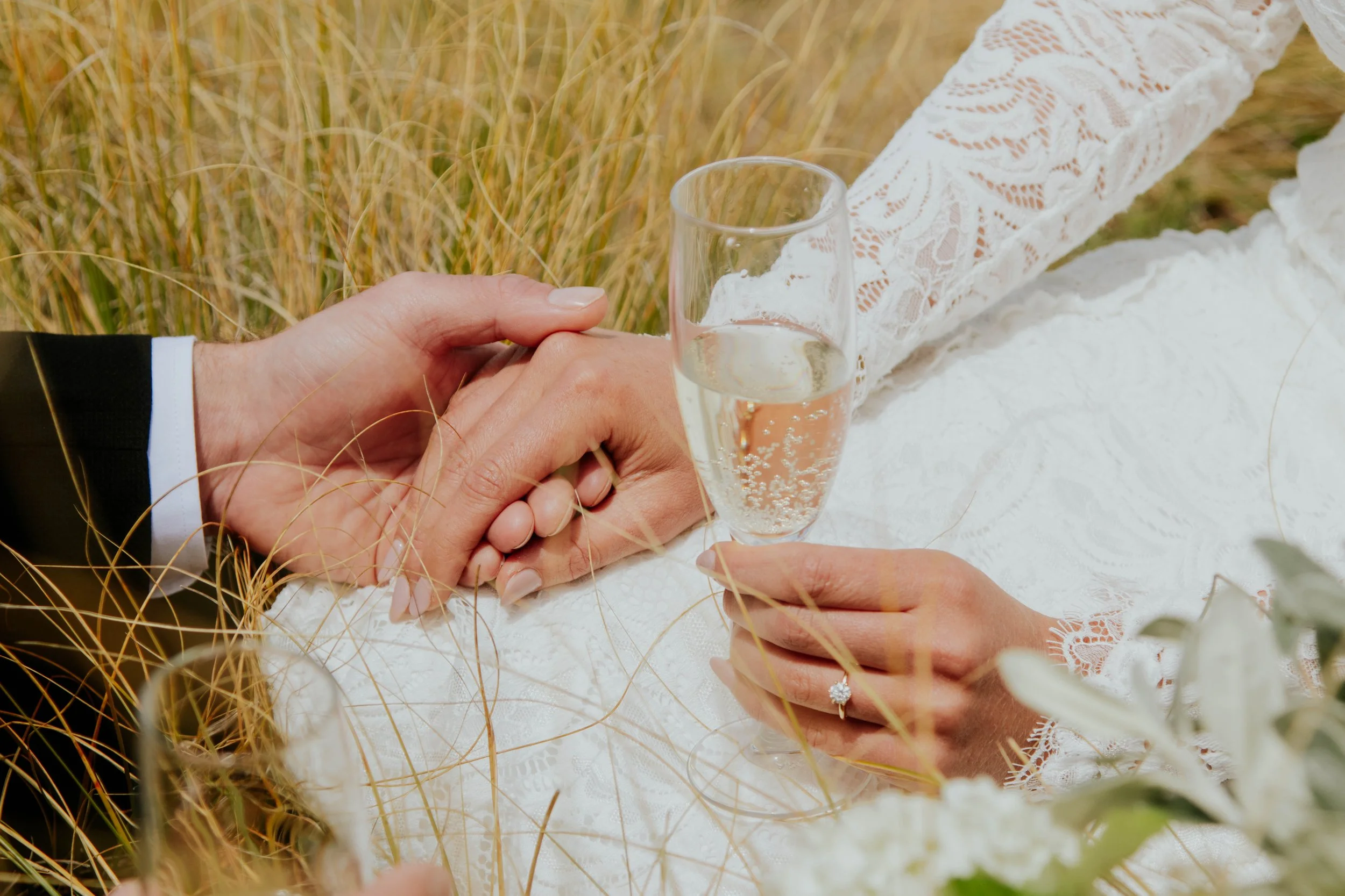 Close-up of a bride and groom holding hands on the ground in a field of tall grass. The bride's hand shows a wedding ring and she holds a glass of champagne. The groom's hand is gently holding hers.