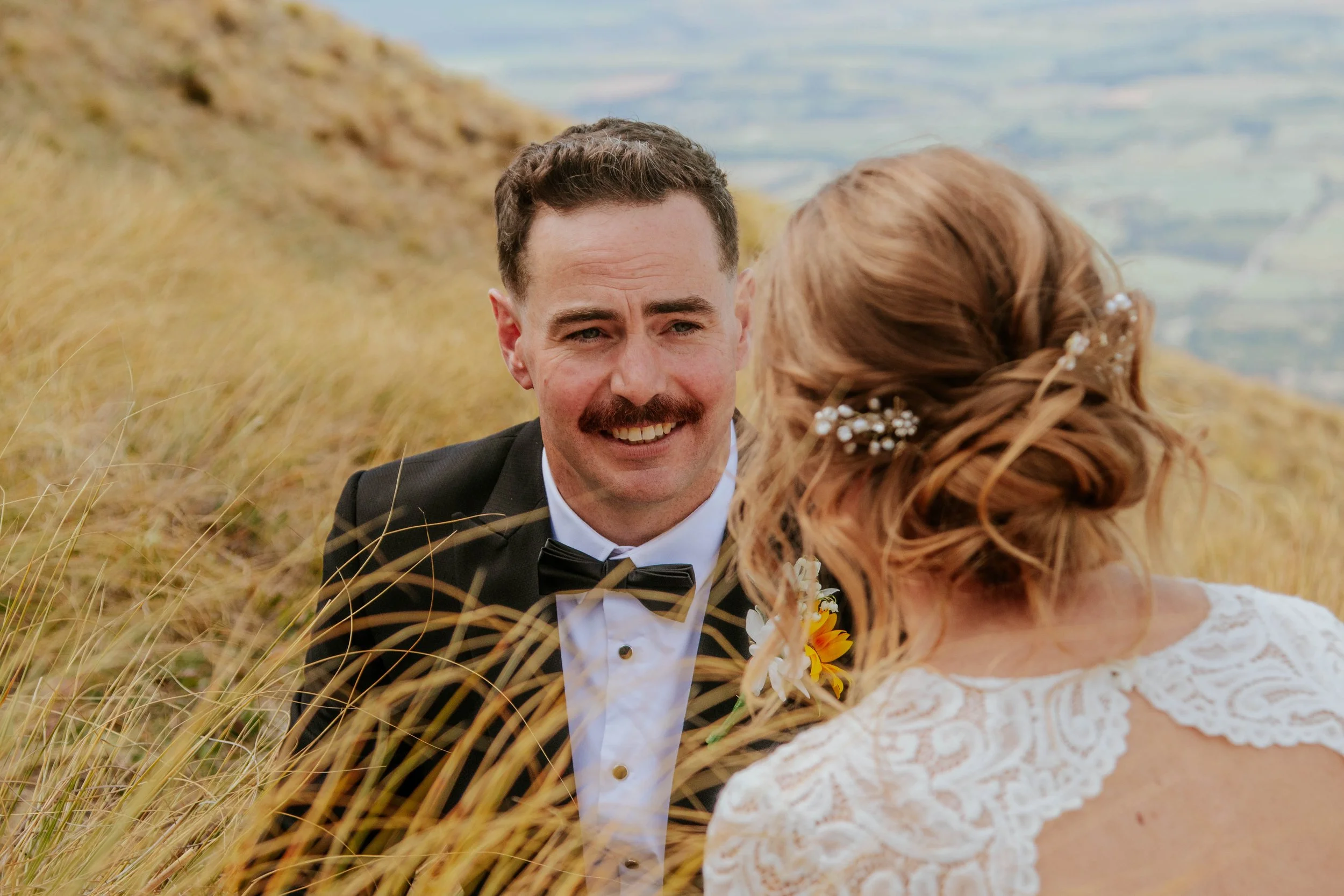 A man in a tuxedo with a bow tie and a woman in a white lace dress are looking at each other in a grassy outdoor setting, possibly during a wedding.
