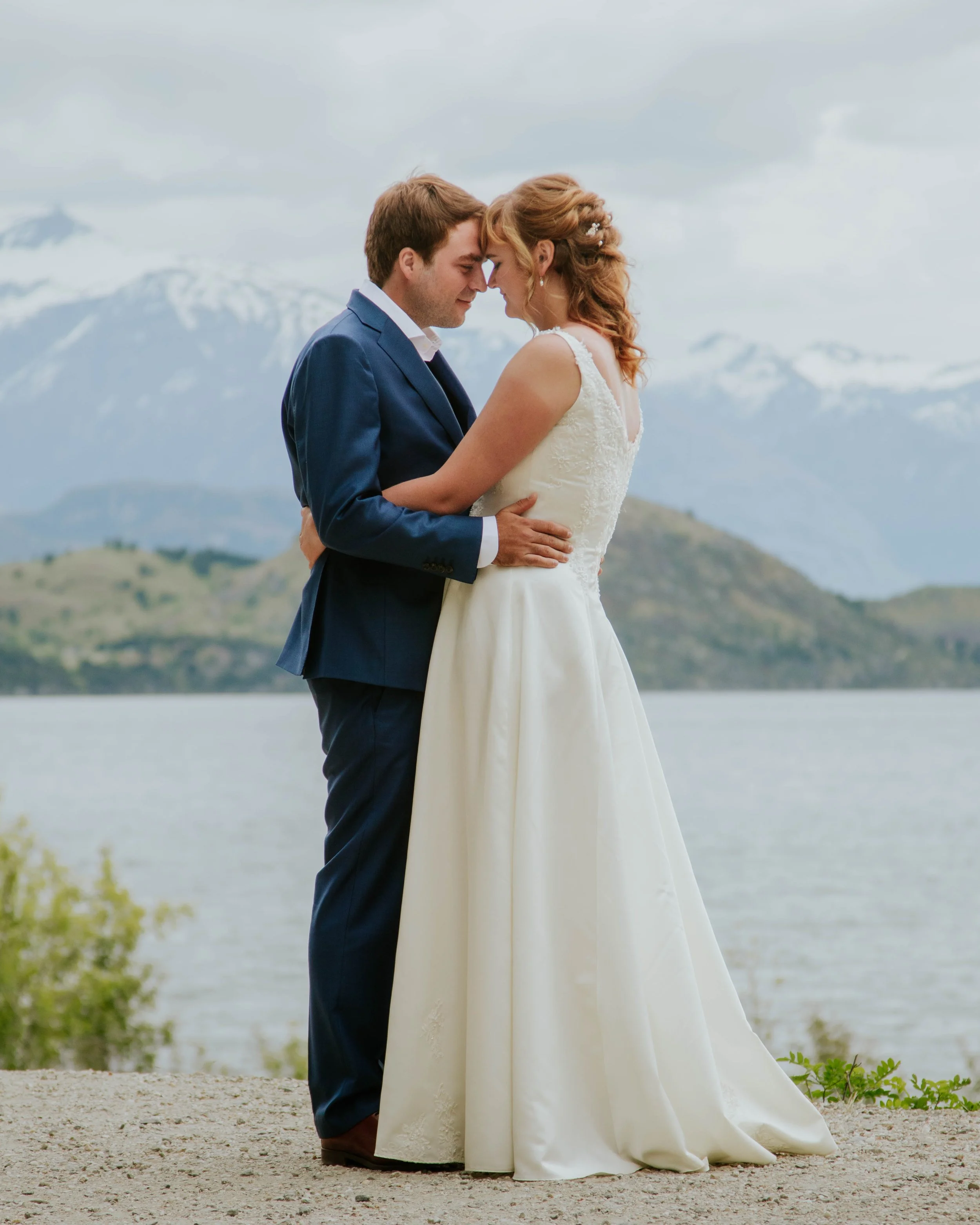 A bride and groom stand close together with foreheads touching during their outdoor wedding, with mountains and a lake in the background.