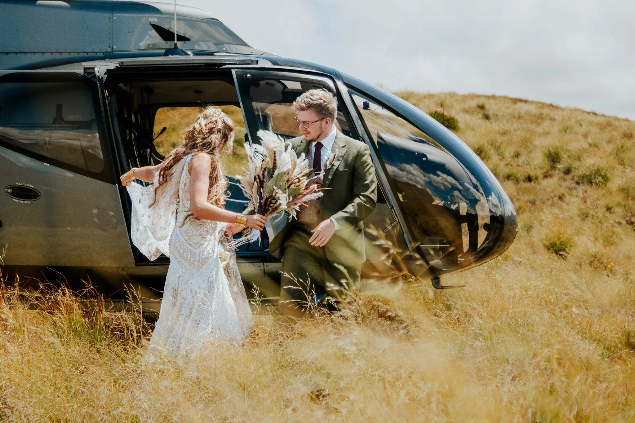 A bride and groom with light skin exchanging vows near a helicopter in a grassy field with hills in the background.