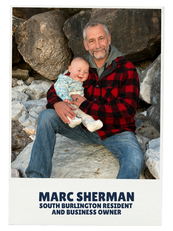 A man with gray hair and a beard, wearing a red and black plaid jacket, sitting on rocks outdoors, holding a smiling baby girl dressed in light-colored clothing with a blue patterned sweater. The background consists of large rocks and stones.