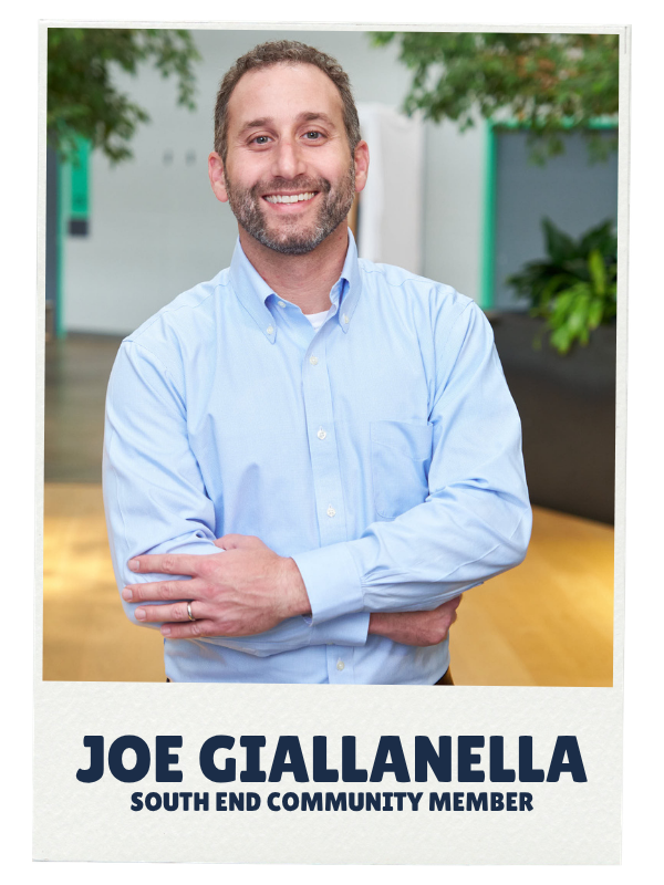 A smiling man with a beard wearing a light blue button-up shirt standing indoors with arms crossed, with green plants and a blurred background. Caption reads "Joe Giallella."