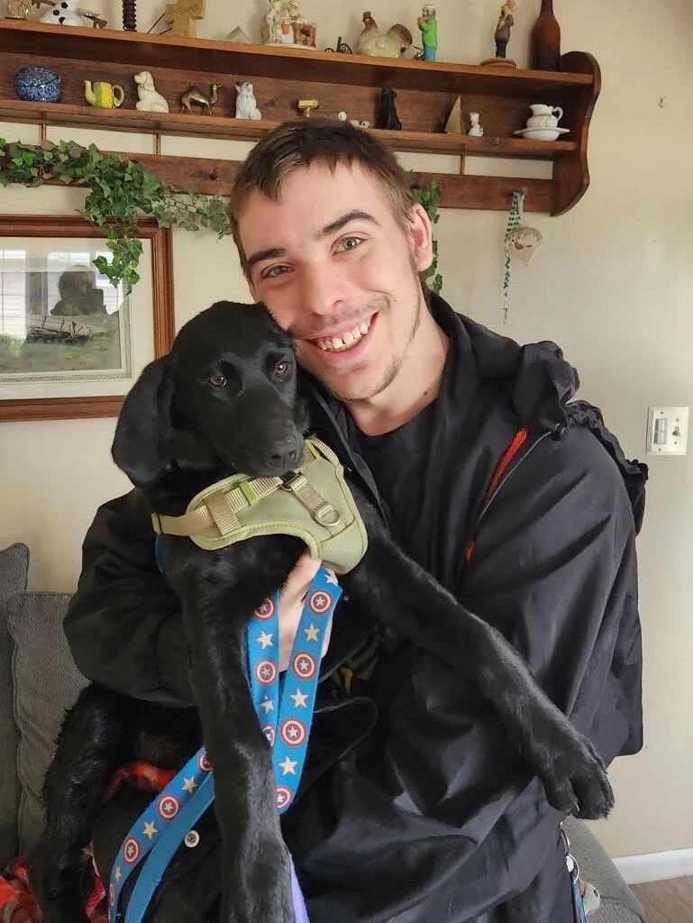 A young man with short brown hair and a beard smiling while holding a black puppy with floppy ears, wearing a harness and a dog-related leash, in a home setting with shelves and decorations in the background.