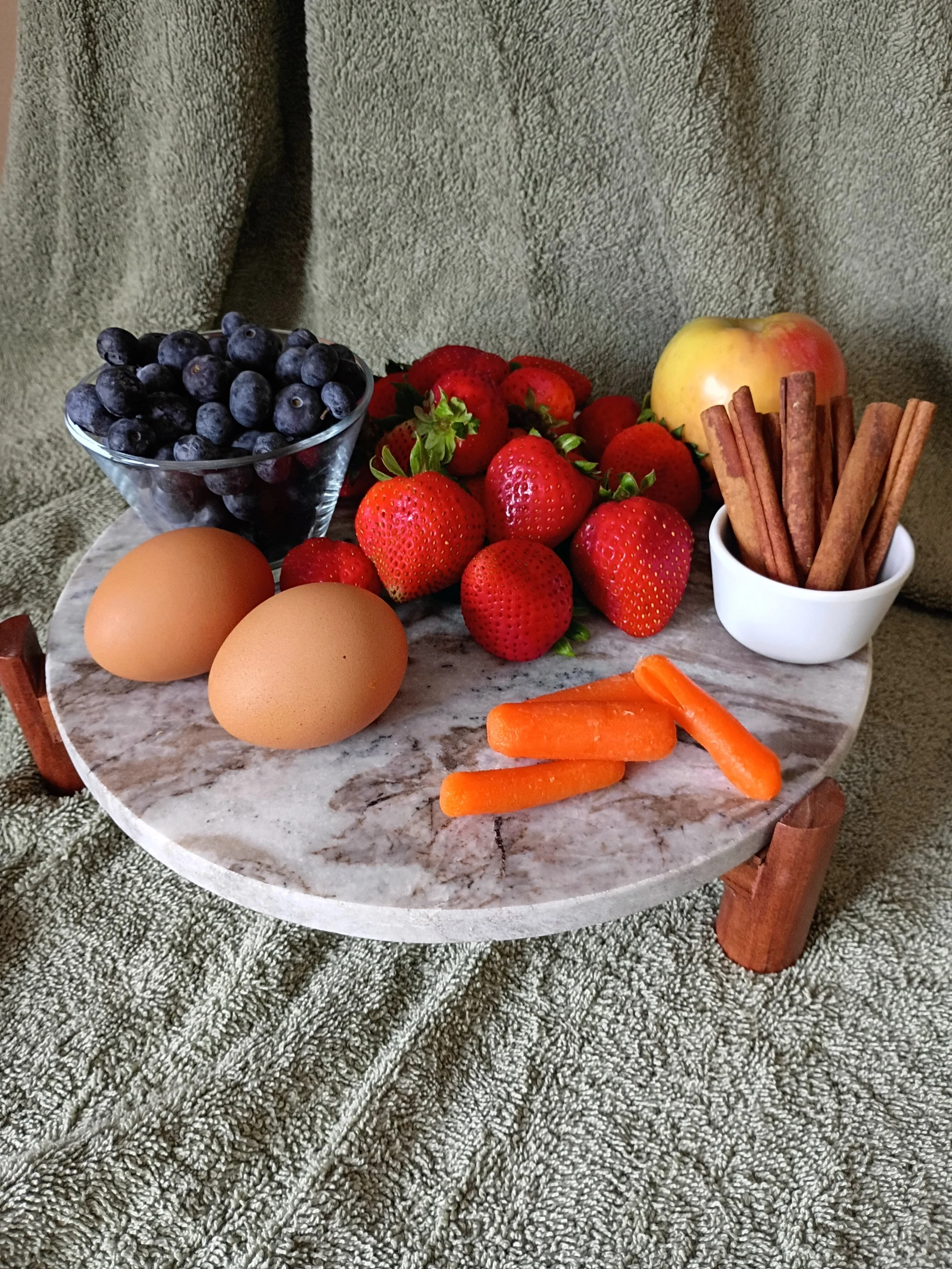 Assorted berries, eggs, carrots, apple, and cinnamon sticks on a marble table, with a grey textured background.