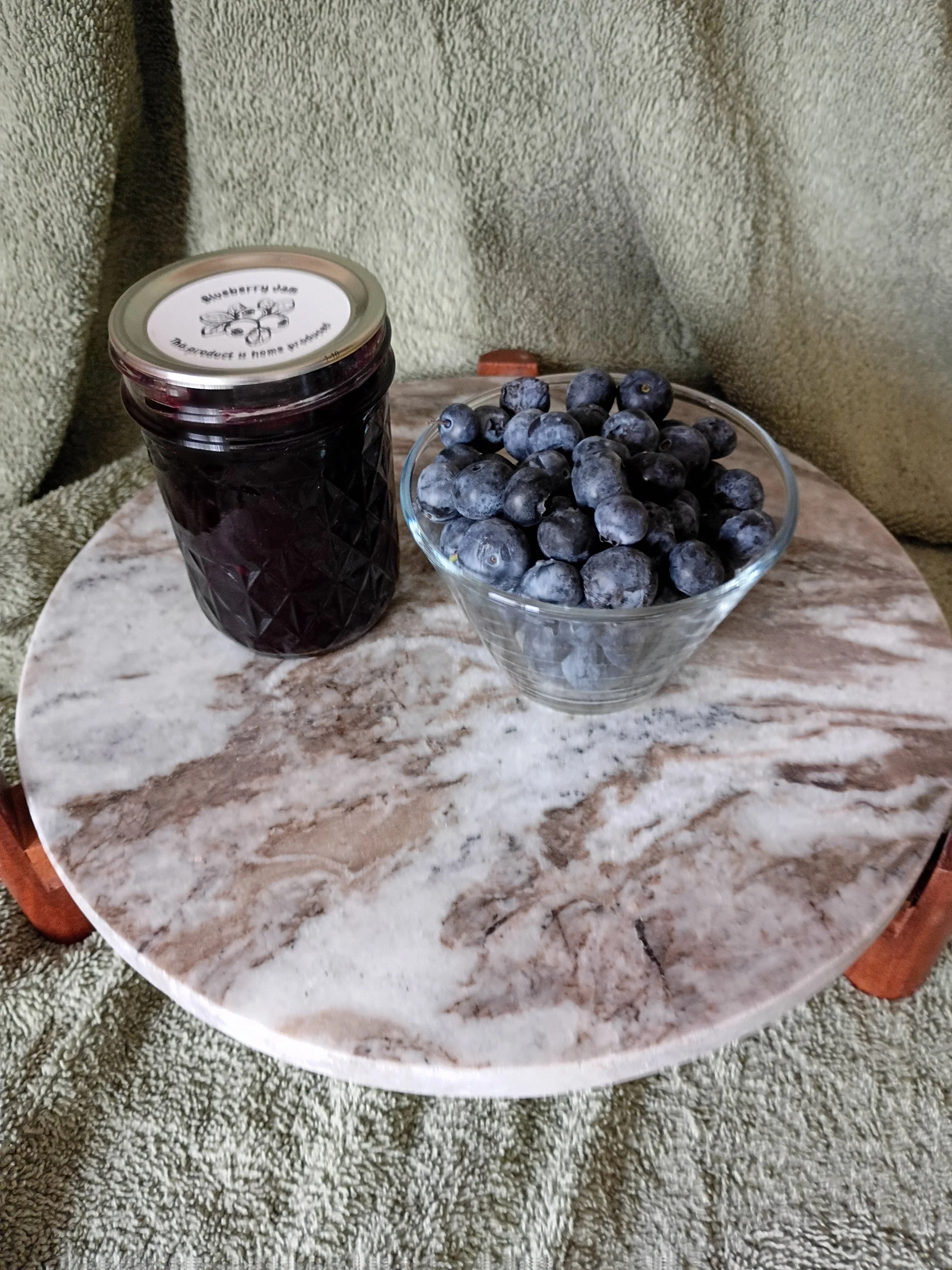A glass bowl filled with blueberries and a jar of blueberry jam on a marble table, with green textured fabric in the background.