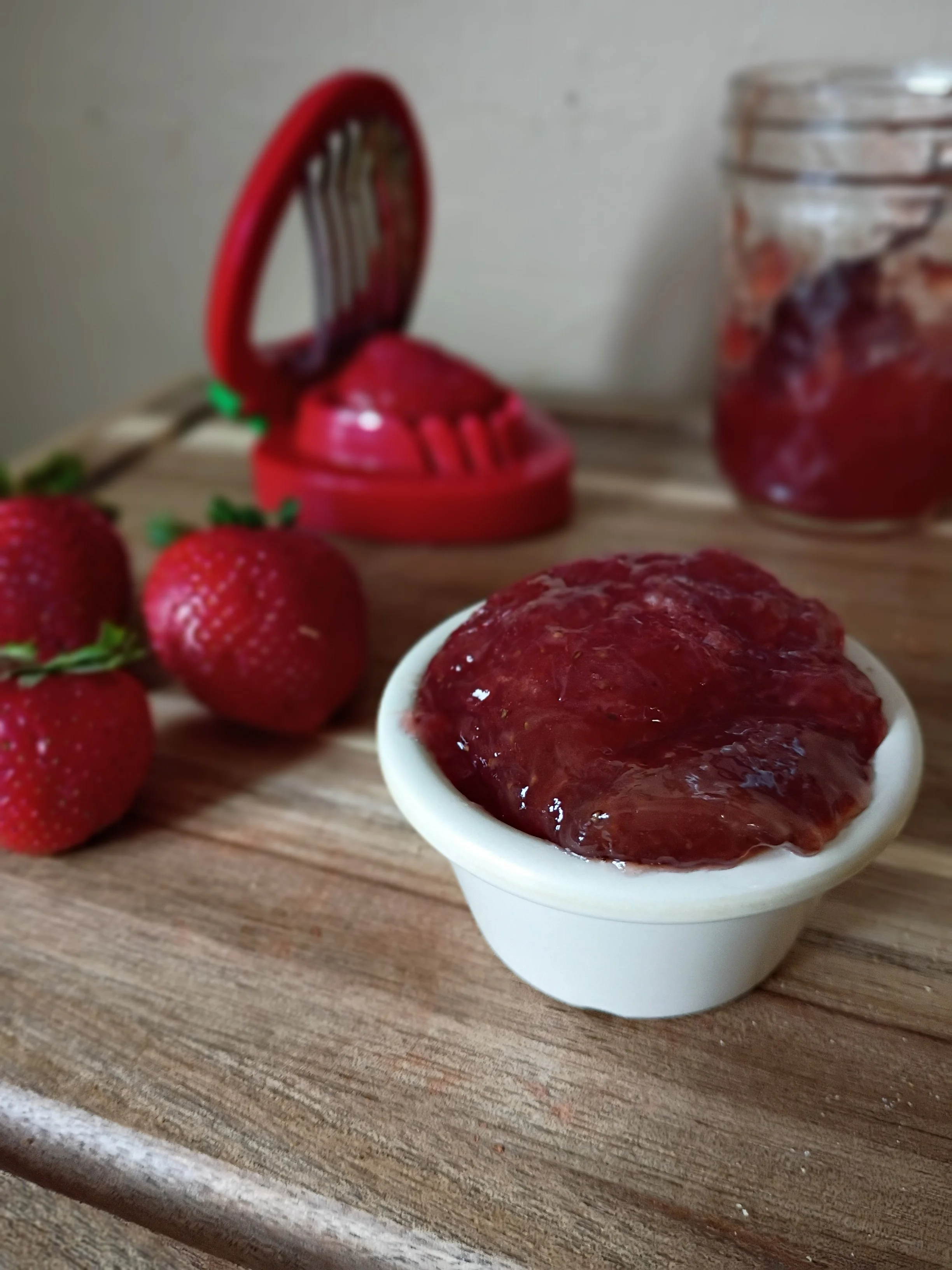 Close-up of a bowl of strawberry jam on a wooden surface, with fresh strawberries and a jar of jam in the background.
