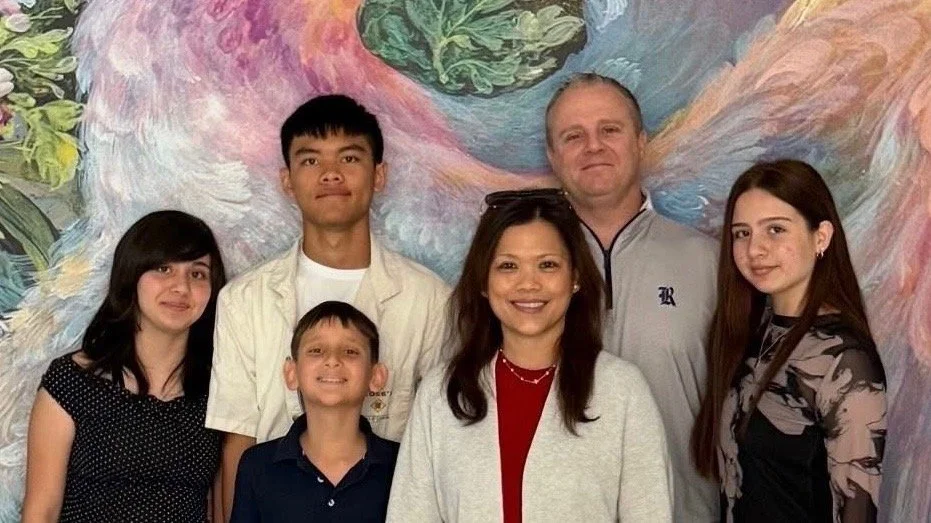 A group of seven people, including two men, one woman, and four children, standing in front of a colorful abstract mural.