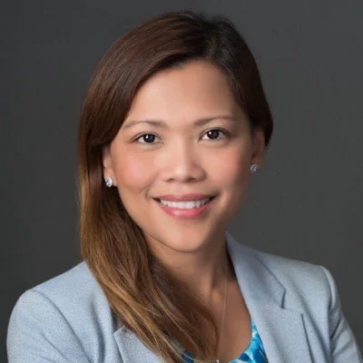 Portrait of a smiling woman with long brown hair, wearing a light gray blazer and a blue patterned blouse, against a gray background.
