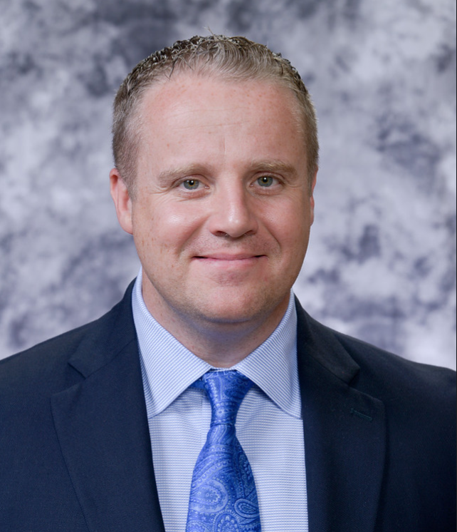 A professional headshot of a man with short blonde hair, wearing a navy suit, light blue shirt, and a blue paisley tie, smiling in front of a grey and white mottled background.
