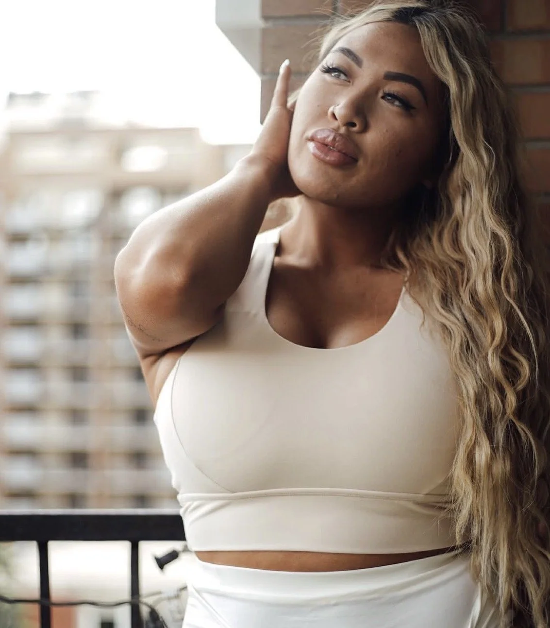 A woman with long, wavy blonde hair standing on a balcony, looking thoughtfully into the distance with her hand touching her head.