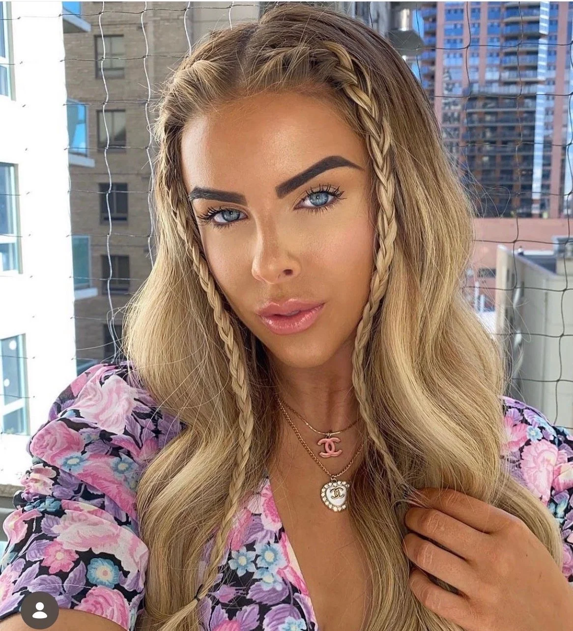 A woman with long blonde hair and blue eyes taking a selfie on a balcony with city buildings in the background. She is wearing a floral top and layered necklaces with logo pendants.
