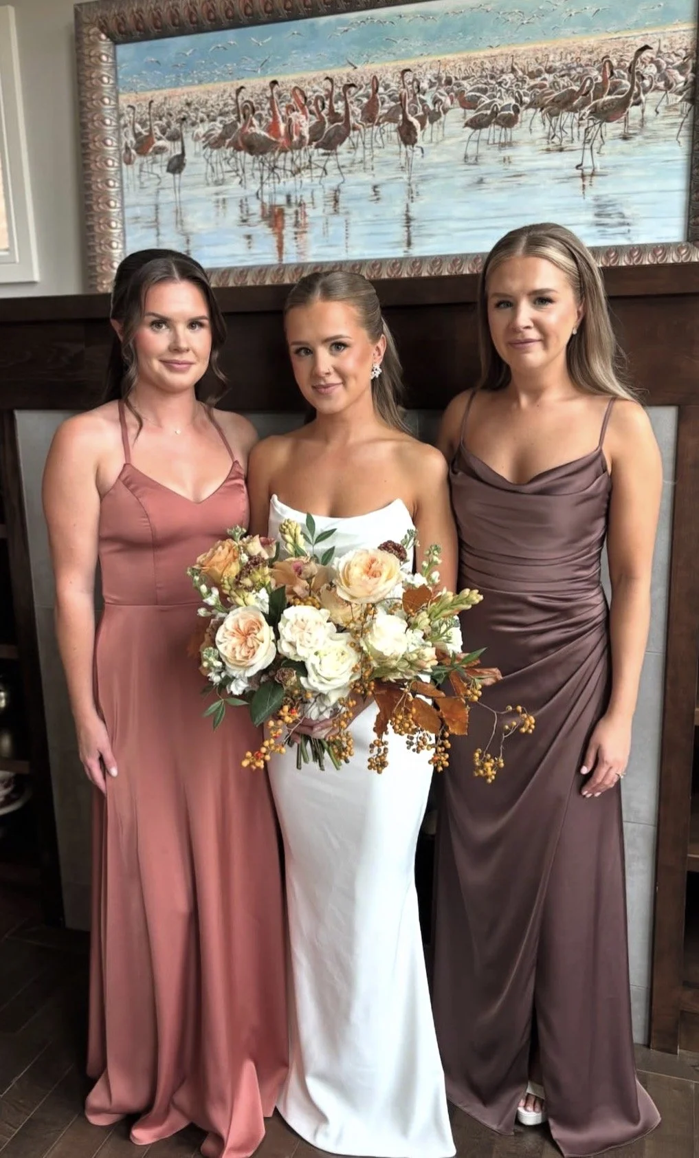 Three women in gown and bridesmaid dresses posing in front of a painting of flamingos on a beach, with the bride in the center holding a large bouquet of flowers.