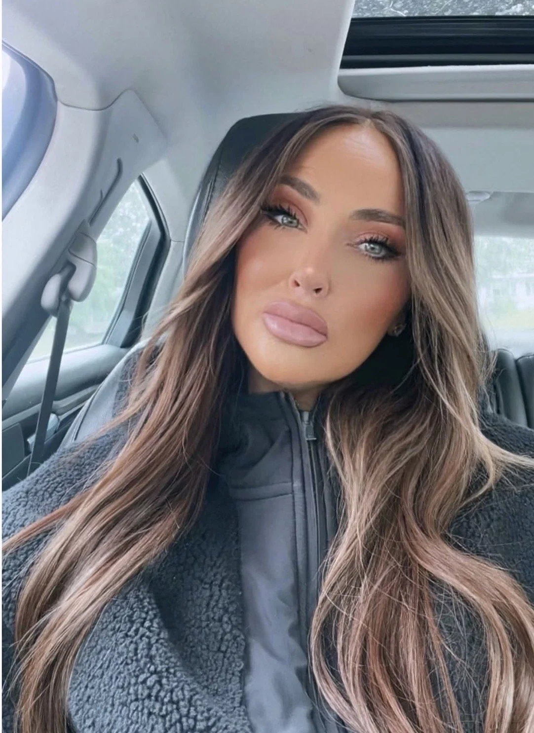 A woman with long, wavy brown hair and blue eyes sitting in a vehicle, wearing a black zip-up jacket.