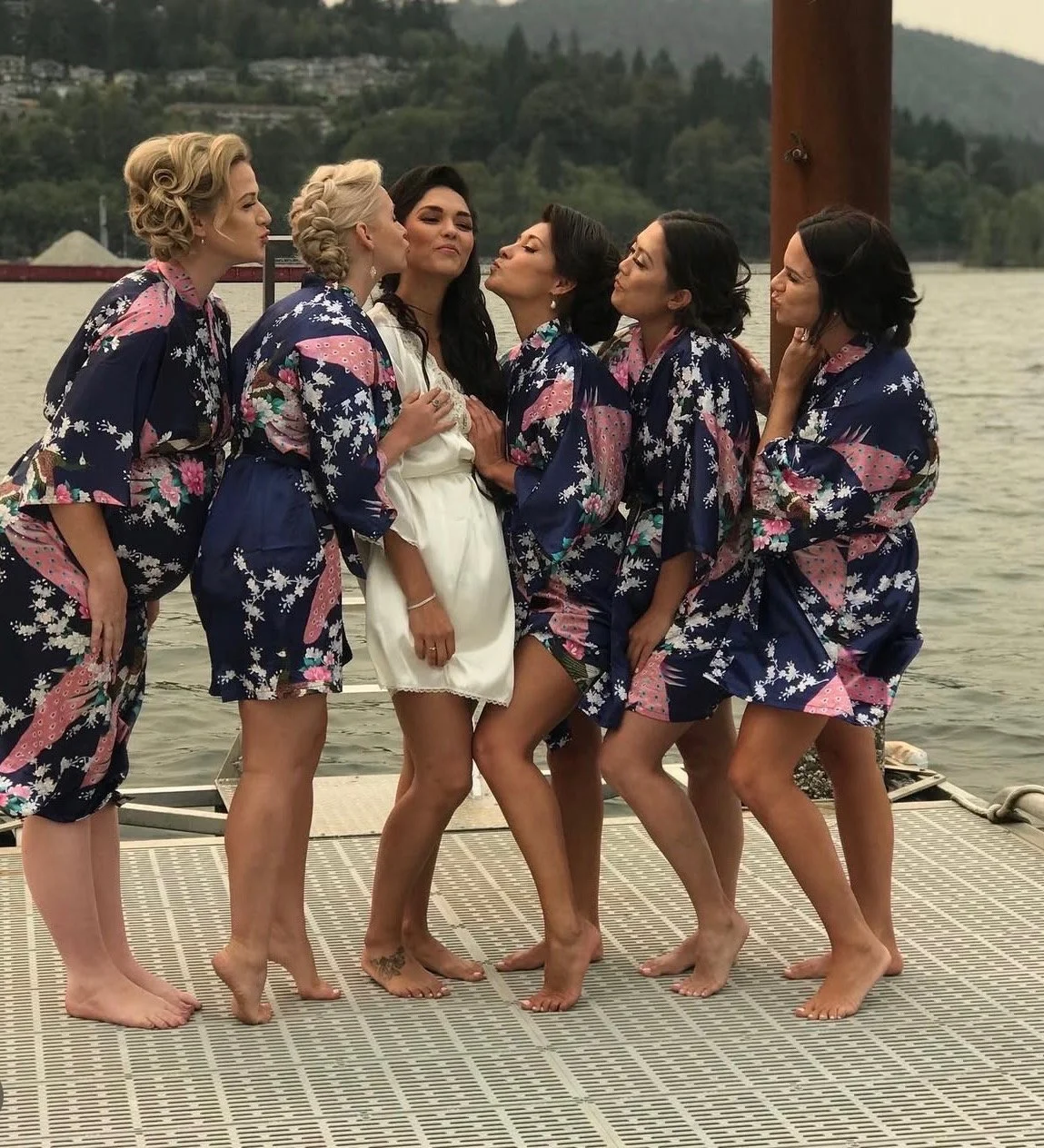 Group of women on a dock near water, posing for a photo with some kissing and others making playful expressions, all wearing matching floral robes, during daytime.