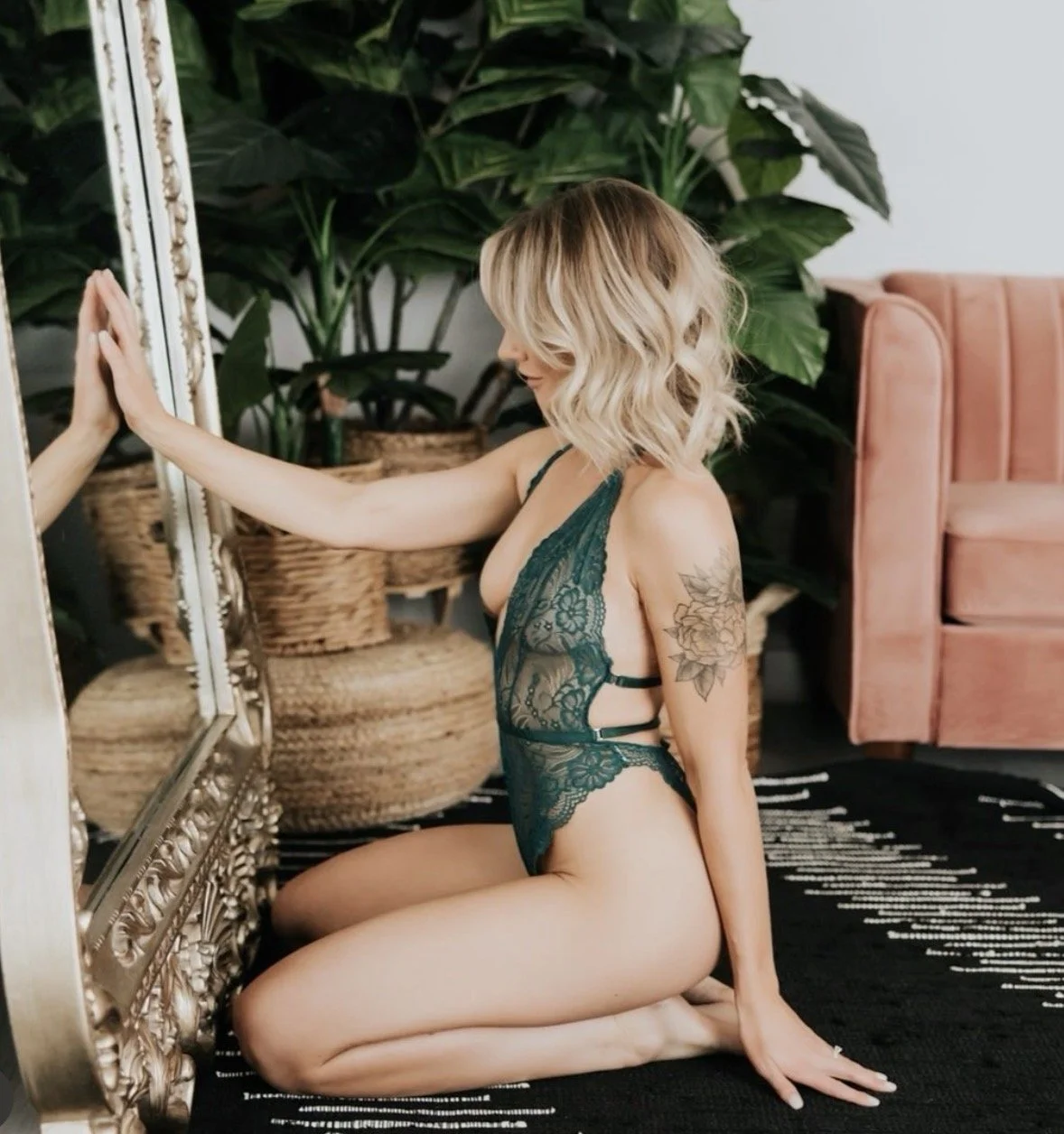 Woman with blonde wavy hair wearing teal lace lingerie, kneeling on a black rug, touching a large ornate mirror, with green plants and pink couch in background.