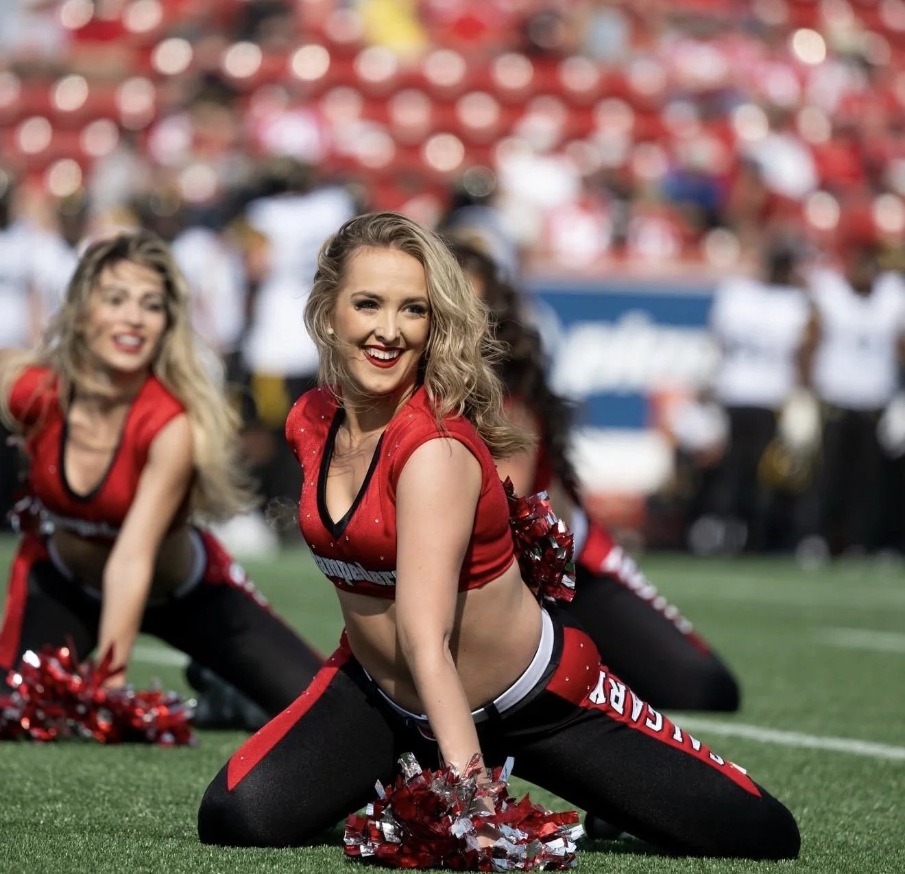Cheerleaders performing a dance routine on a football field during a game, wearing red and black uniforms with pom-poms.