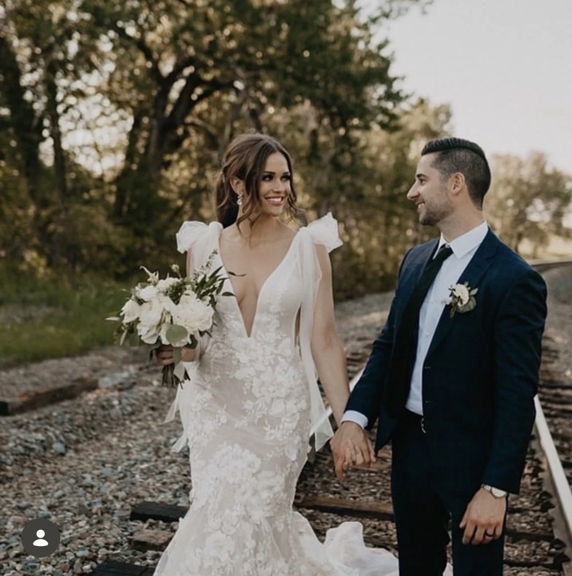 A bride and groom holding hands outdoors near train tracks, with trees and a sunset in the background, celebrating their wedding day.