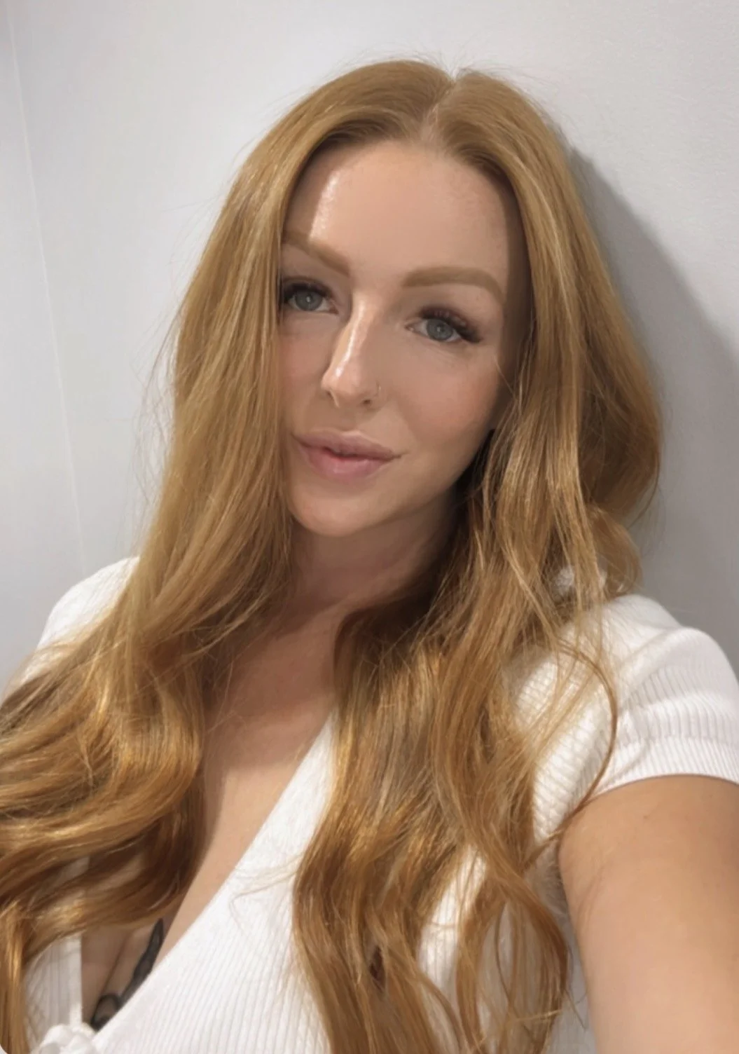 Close-up of a woman with long, wavy red hair, blue eyes, and light makeup, wearing a white top, standing against a plain white wall.