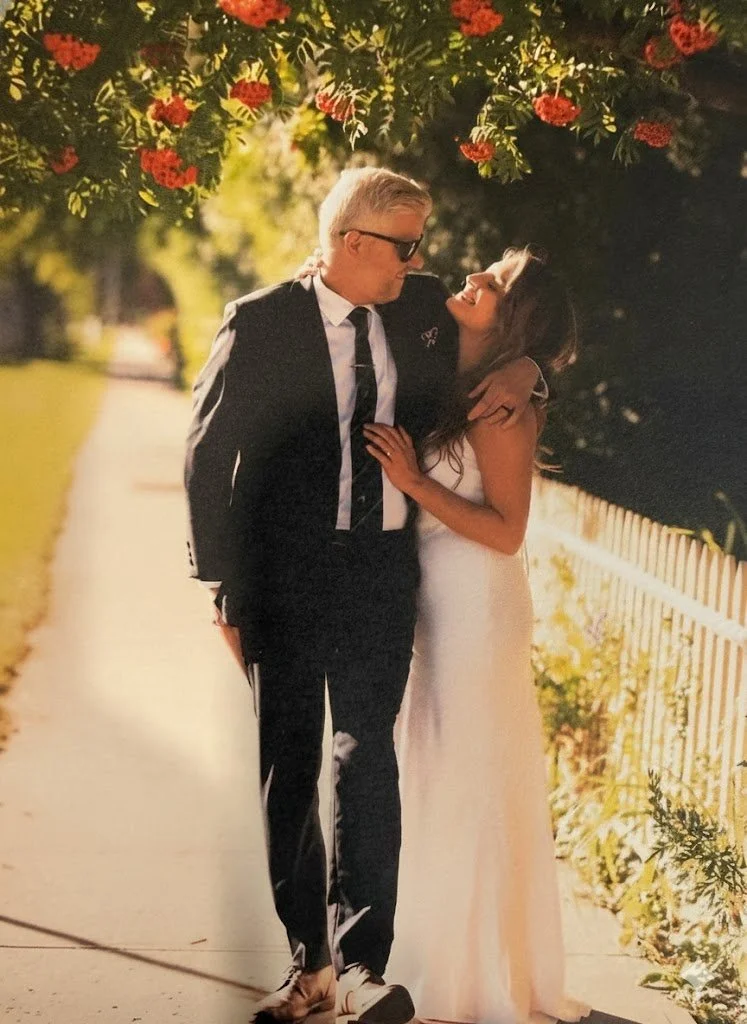 A man in a black suit and sunglasses stands close to a woman in a white dress, on a sunny outdoor walkway with a yellow fence and flowering bushes overhead.