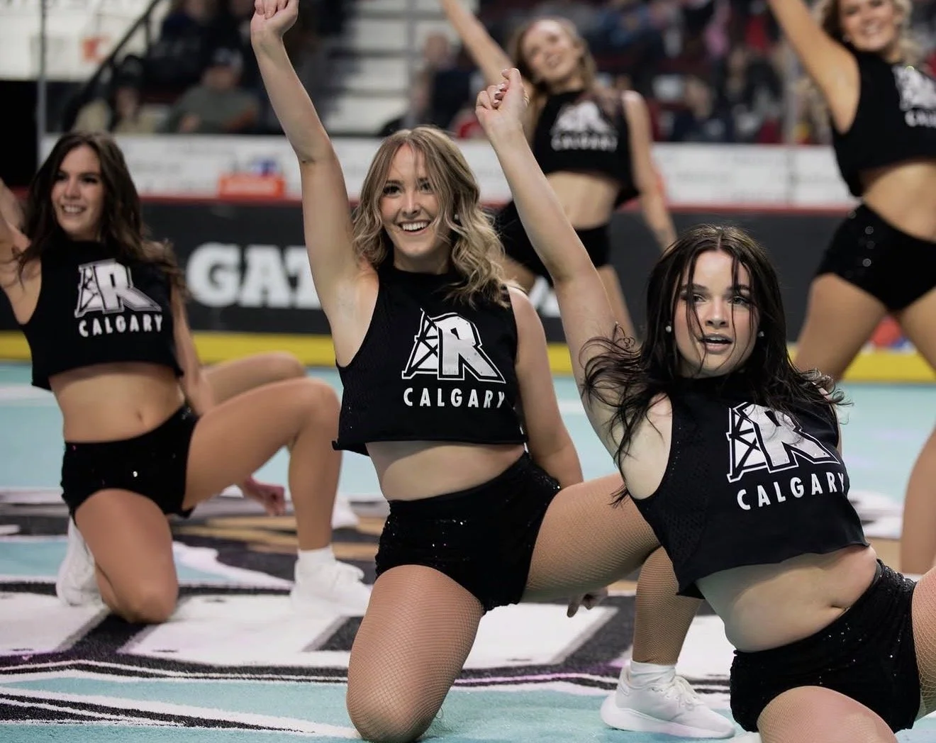 Cheerleaders performing a routine on the field wearing black uniforms with 'Calgary' and a large 'R' logo. The cheerleaders are in various poses, smiling, with some kneeling and others in different stances.