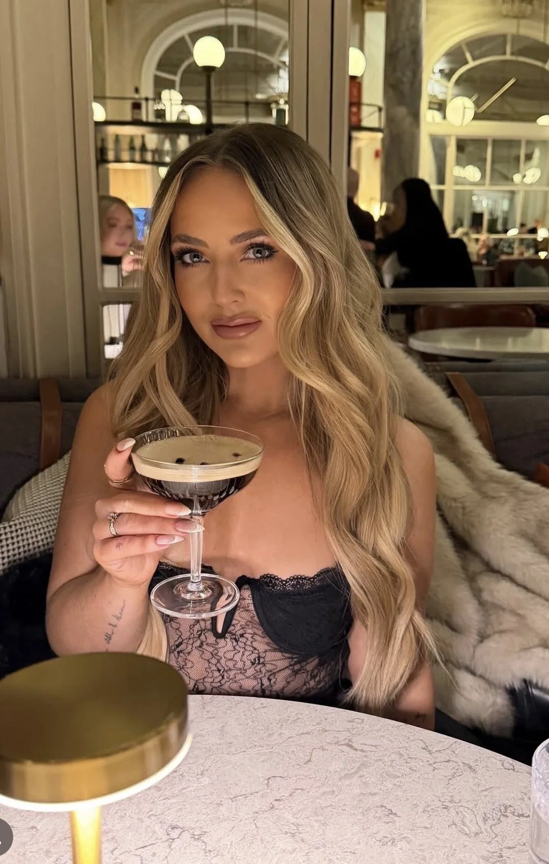A young woman with long wavy blonde hair, holding a cocktail glass filled with a dark-colored drink, sitting at a restaurant table. She is wearing a black lace top and has a light-colored fur coat draped over her shoulder. The background shows a well