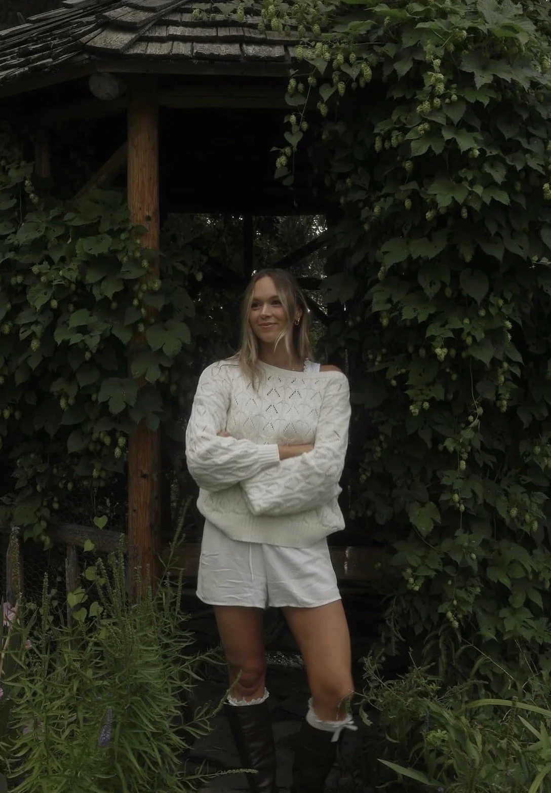 A young woman standing with arms crossed in front of a lush green garden with large foliage and a vine covering a wooden structure.
