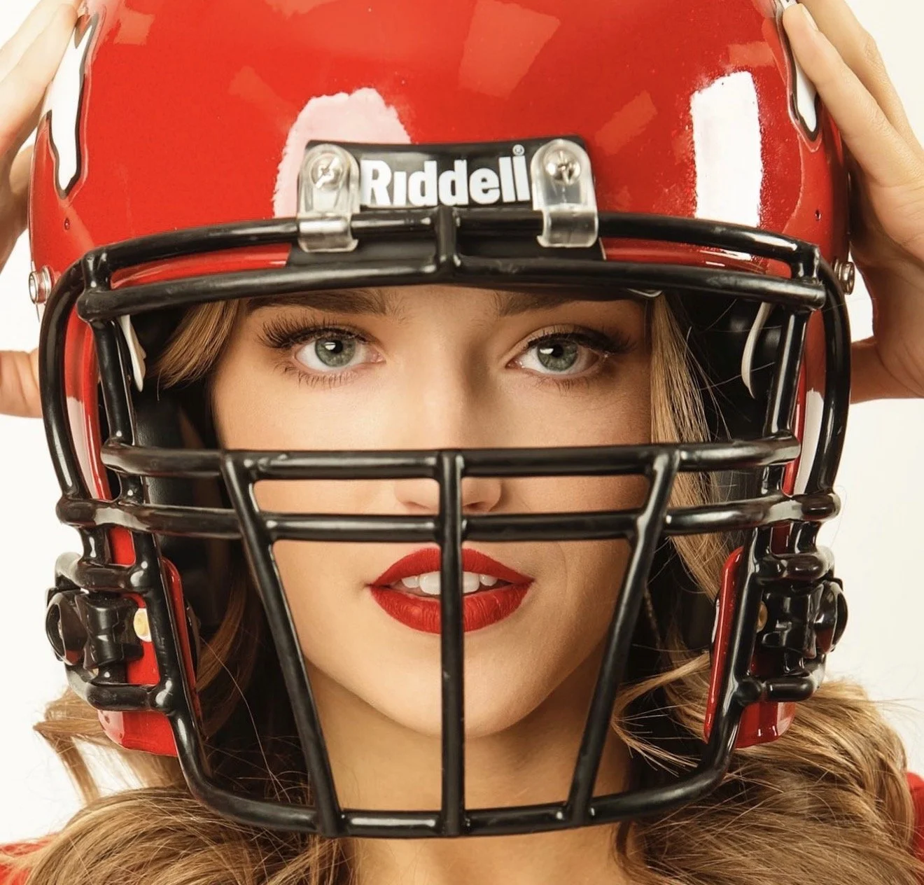 A woman with blue eyes and red lipstick wears a red football helmet, holding it with both hands.