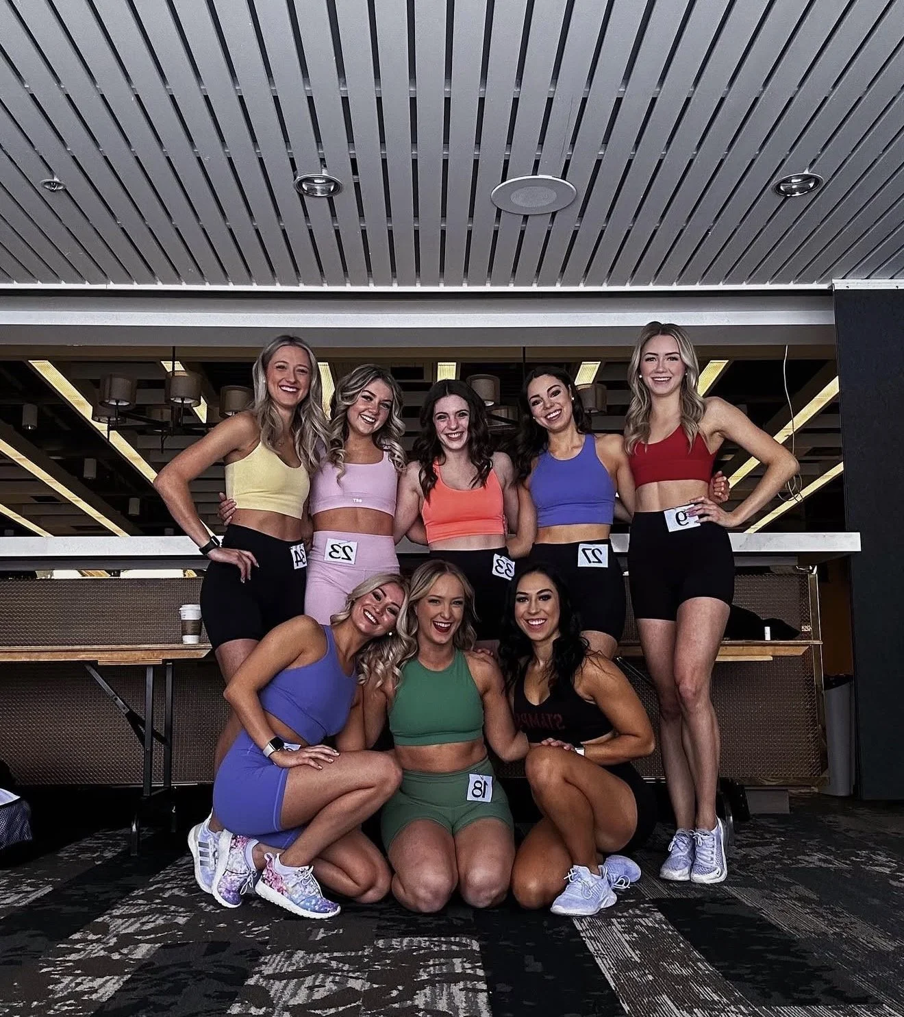 A group of nine women posing together indoors, dressed in colorful athletic outfits with numbers attached, possibly participating in a fitness or bodybuilding competition.