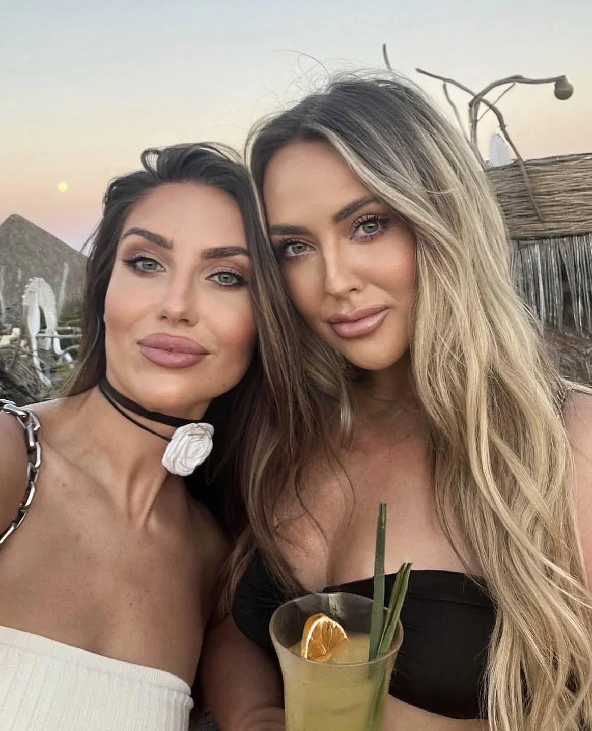 Two women with long hair and makeup pose for a selfie outdoors during sunset, with a drink containing orange slice and green straw in front of them.