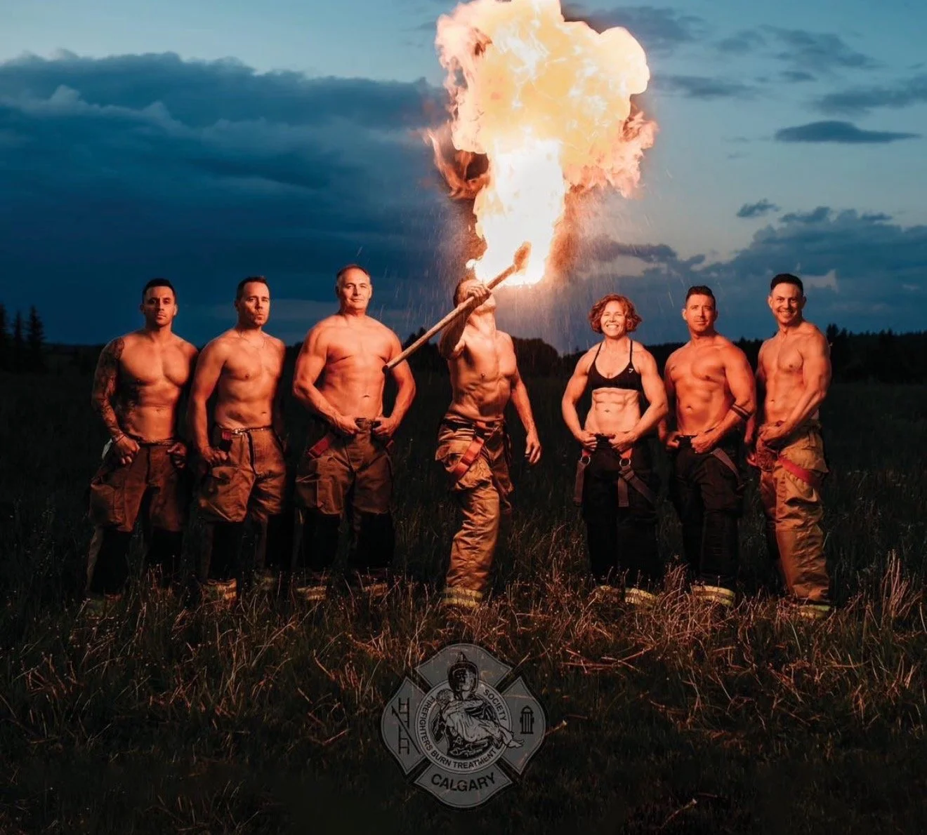 A group of seven shirtless firefighters standing outdoors at dusk, with one firefighter holding a torch that is producing a large fireball. They are wearing firefighting pants, and the background shows a cloudy sky and open field. There is a Calgary 