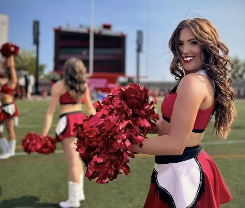 A smiling cheerleader with long, curly brown hair holding red pom-poms on a football field during daytime, with other cheerleaders and a scoreboard in the background.