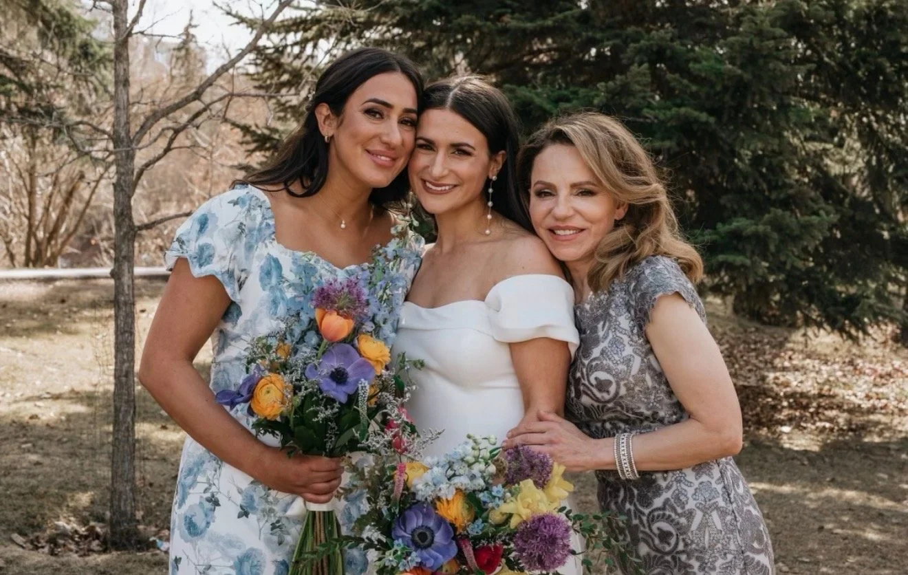 Three women in elegant dresses smiling outdoors with trees in the background, one holding a colorful bouquet of flowers.
