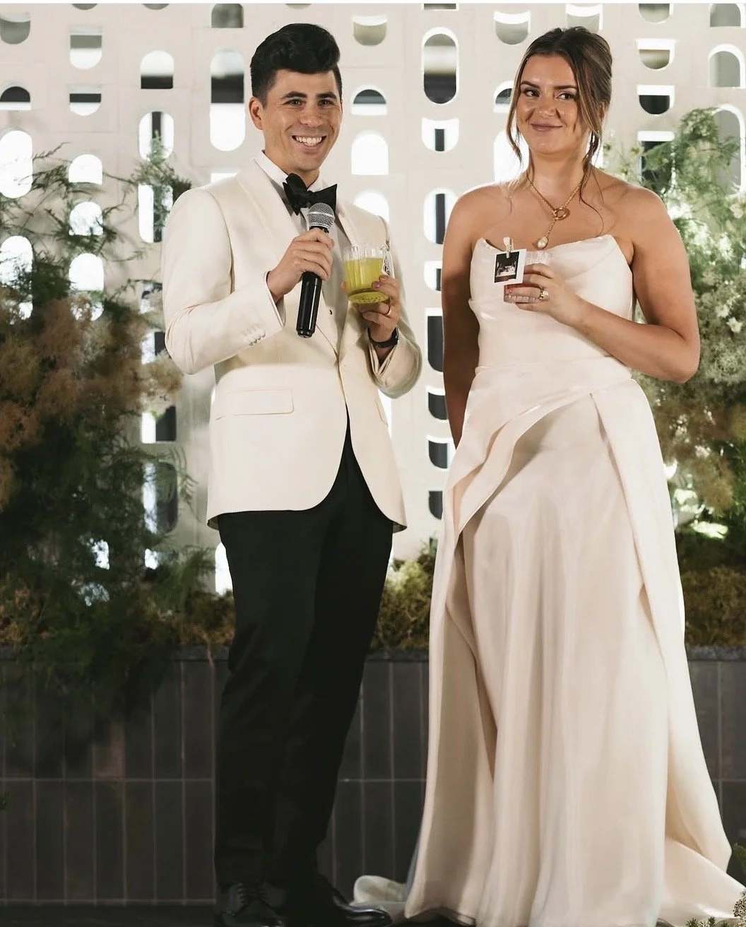 A man and woman in wedding attire standing together at a wedding reception, holding drinks and smiling.