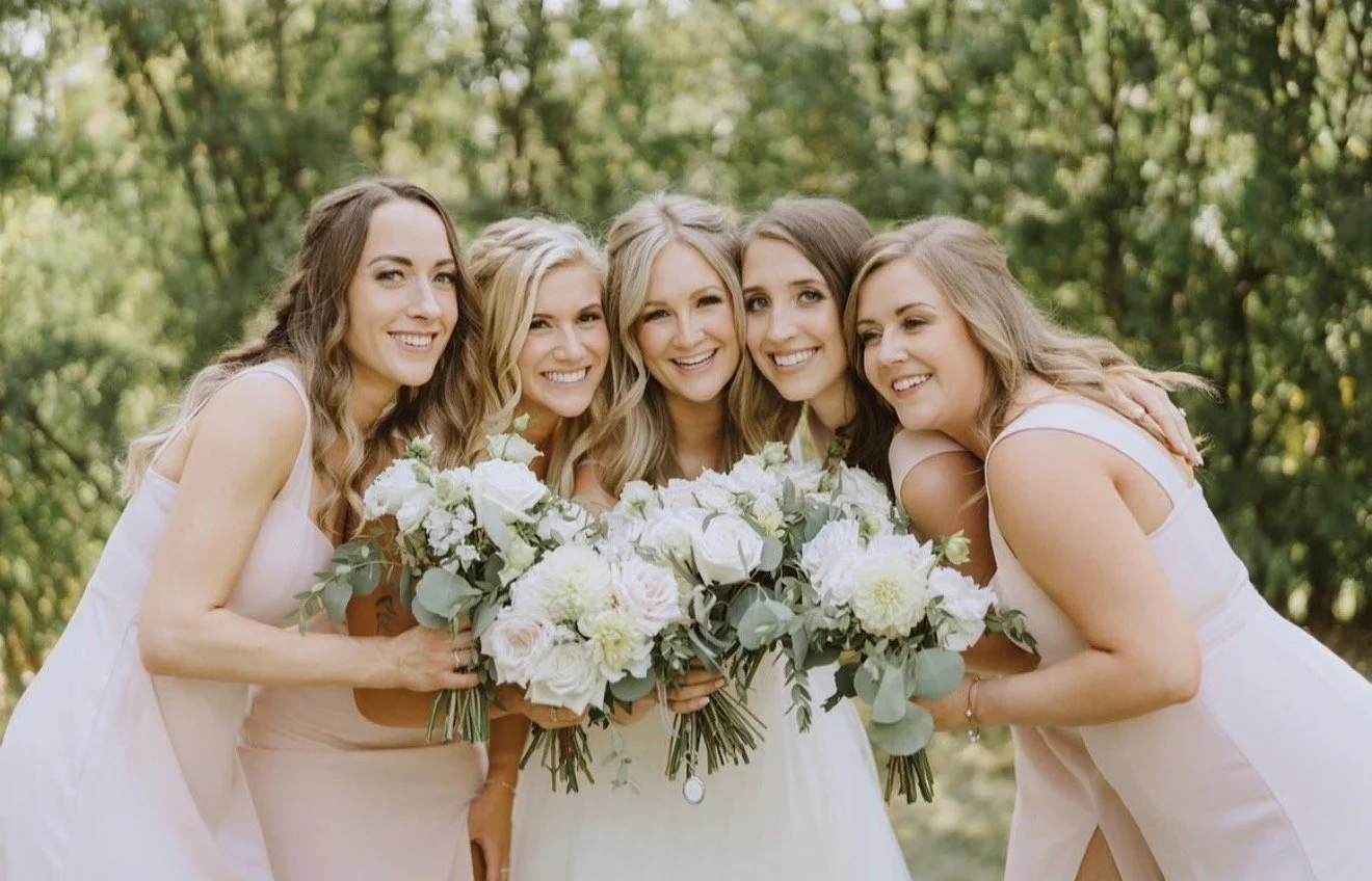 Six women dressed in light-colored dresses standing closely together outdoors, holding large bouquets of white and light pink flowers, smiling for a group photo in a green, wooded area.