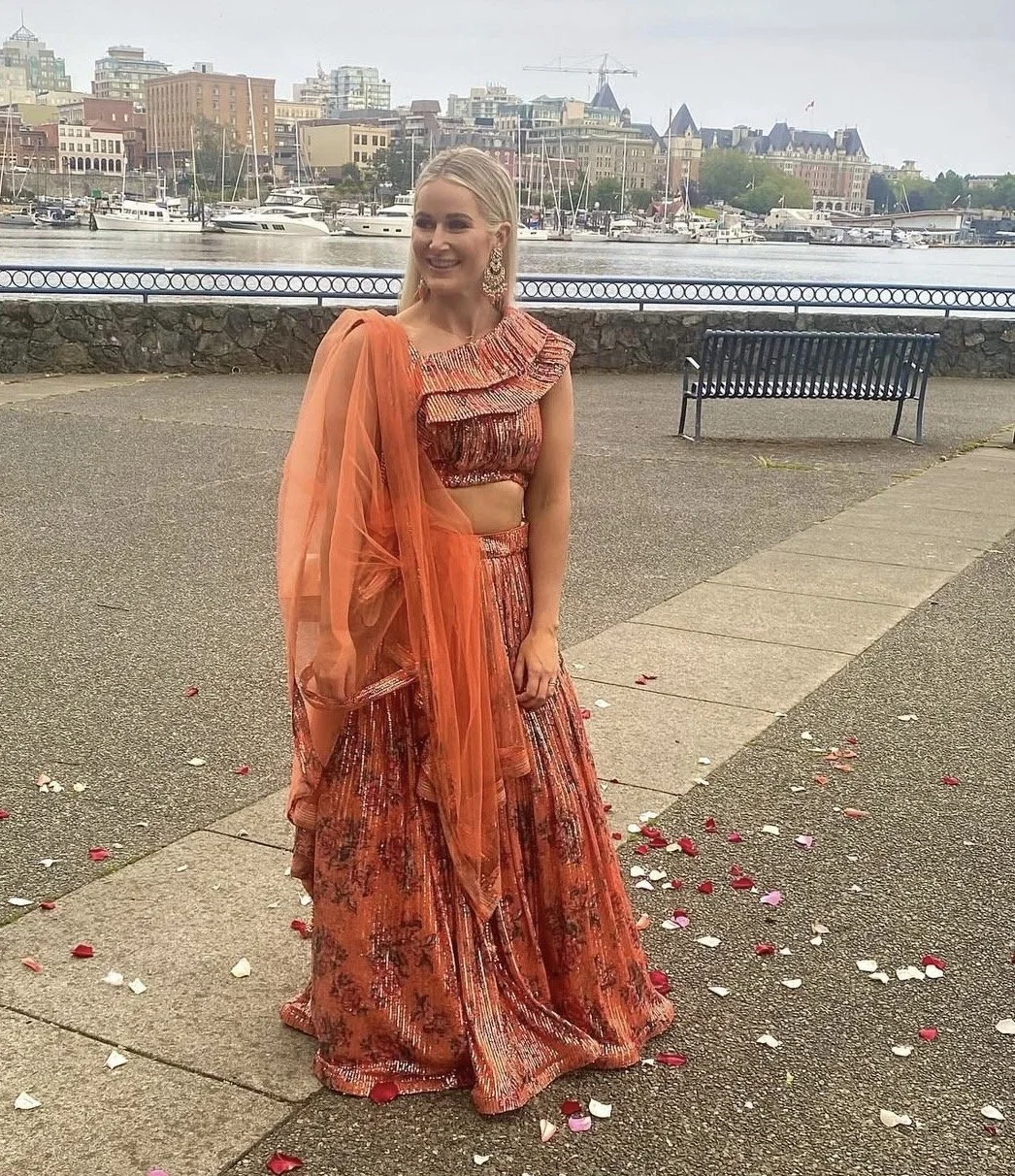 A woman in an orange red traditional dress, standing outdoors near water with boats and buildings in the background, smiling.