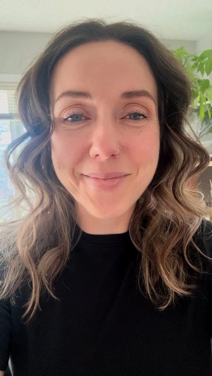 A woman with wavy brown hair and blue eyes smiling, wearing a black top, in a room with window blinds and a green plant in the background.