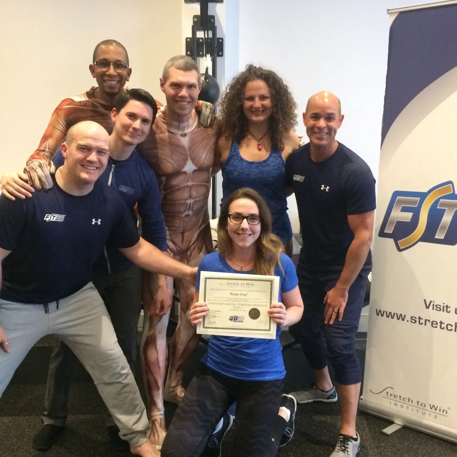 Group of seven adults and one young woman holding a certificate, inside a gym or fitness center with fitness equipment and a banner. The young woman is kneeling in front, holding a certificate for a facial stretch therapy level, surrounded by smiling