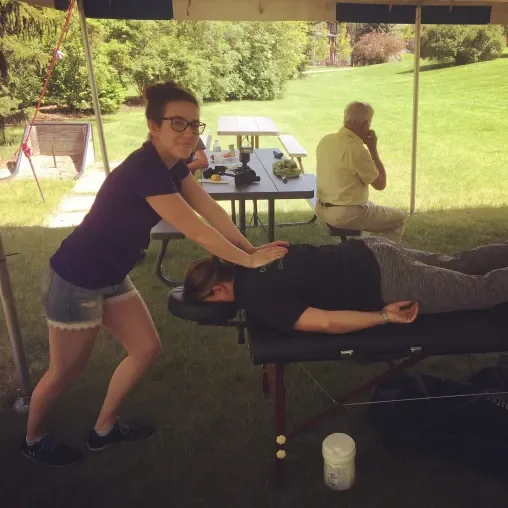 A woman receiving a massage outdoors under a canopy, while a man sits at a picnic table talking on his phone in the background.