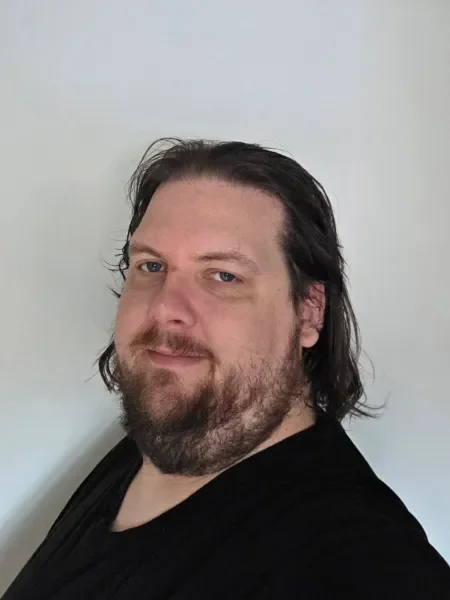 A man with shoulder-length dark hair, a beard, and mustache, wearing a black shirt, standing against a plain white wall.