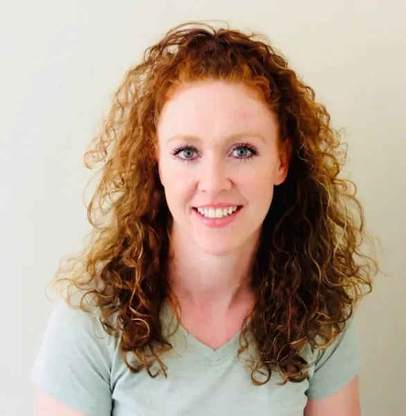 A woman with curly red hair and blue eyes smiling at the camera against a plain, light-colored background.