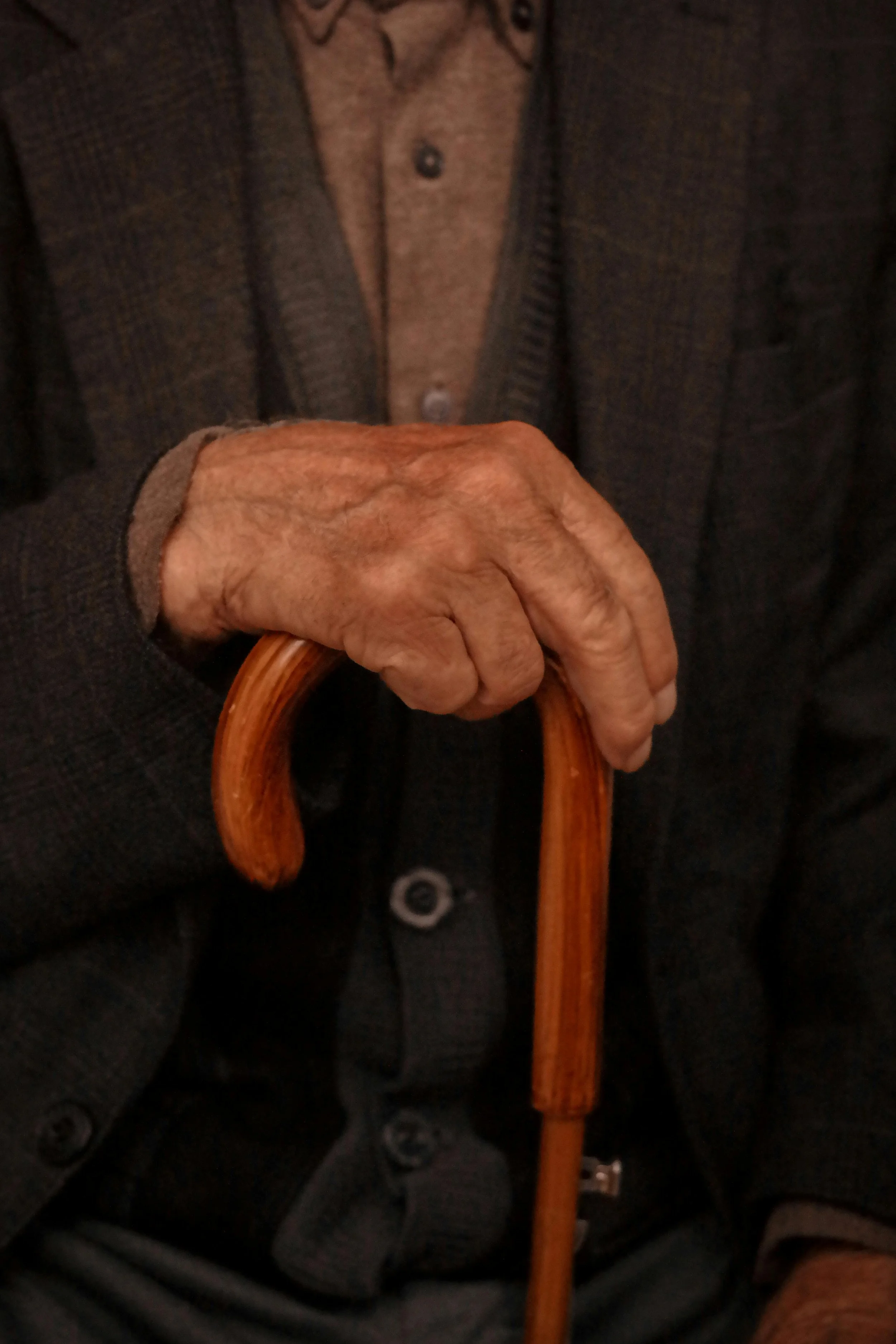 Close-up of an elderly person's hand holding a wooden cane, with the upper part of their body visible, wearing a gray jacket and brown shirt.