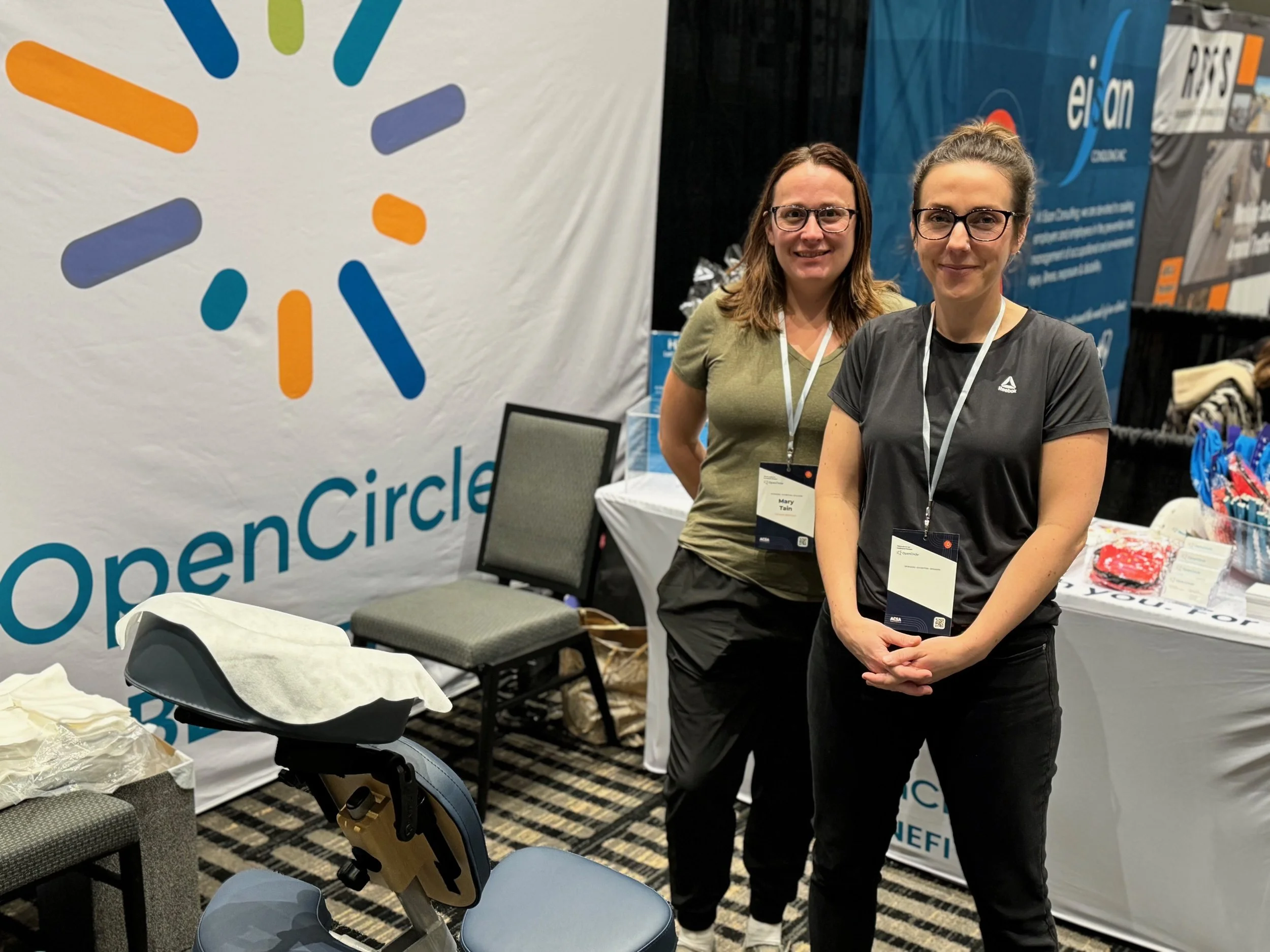 Two women standing in front of a large Open Circle banner at a conference booth. They are smiling and wearing conference badges, with chairs and a table with promotional items behind them.